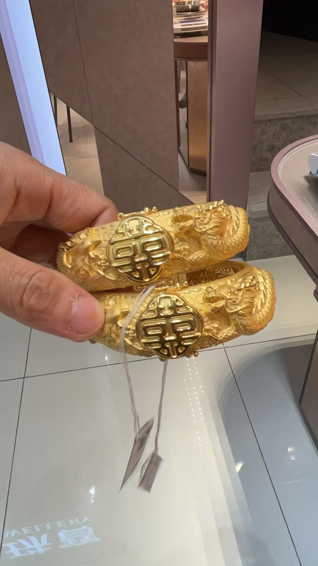 A person holds two ornate, yellow gold dragon-themed bangles featuring a prominent 
