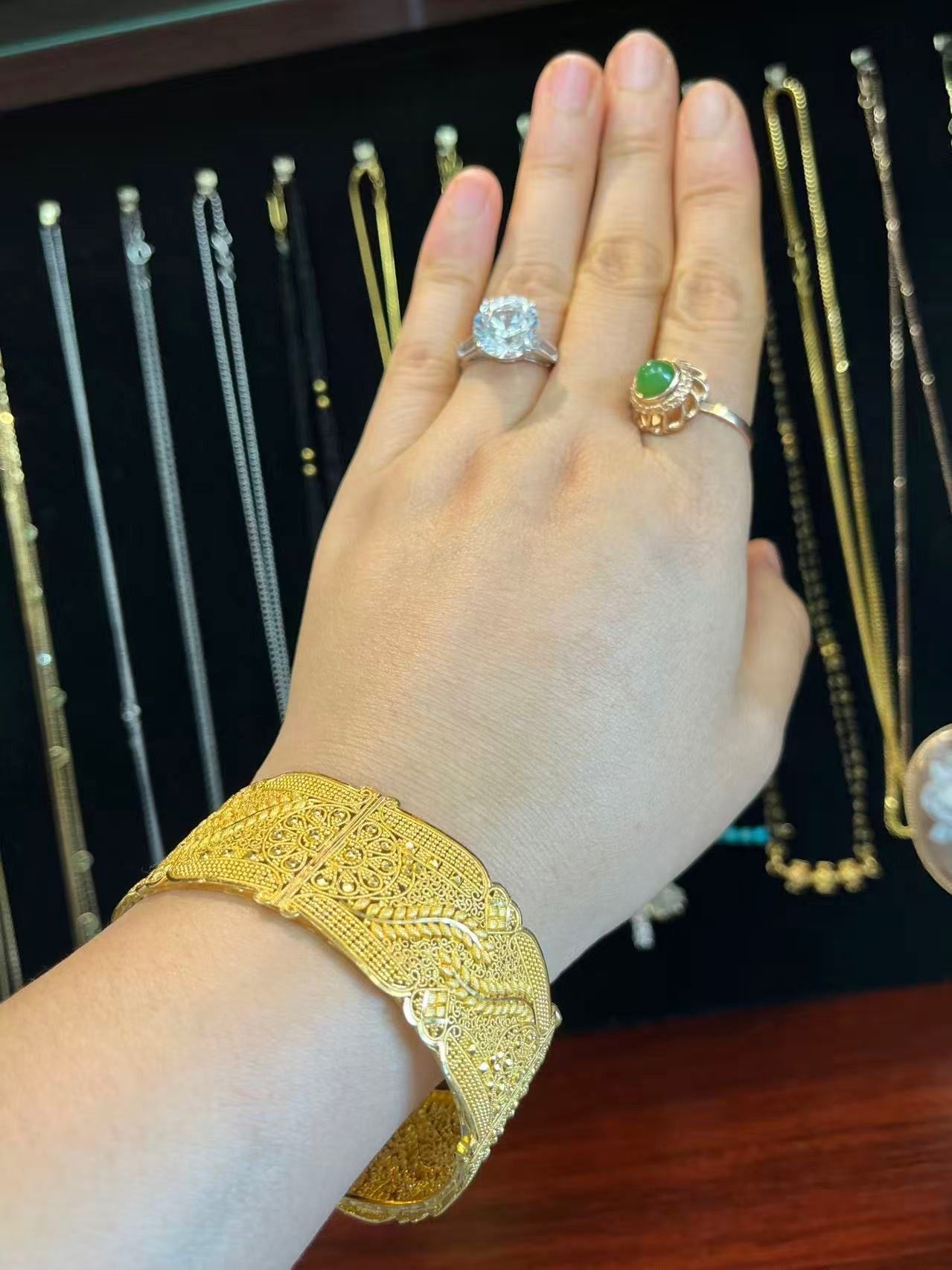 A hand wearing a wide, textured gold cuff bracelet, a diamond ring, and a green jade ring against a backdrop of necklaces.