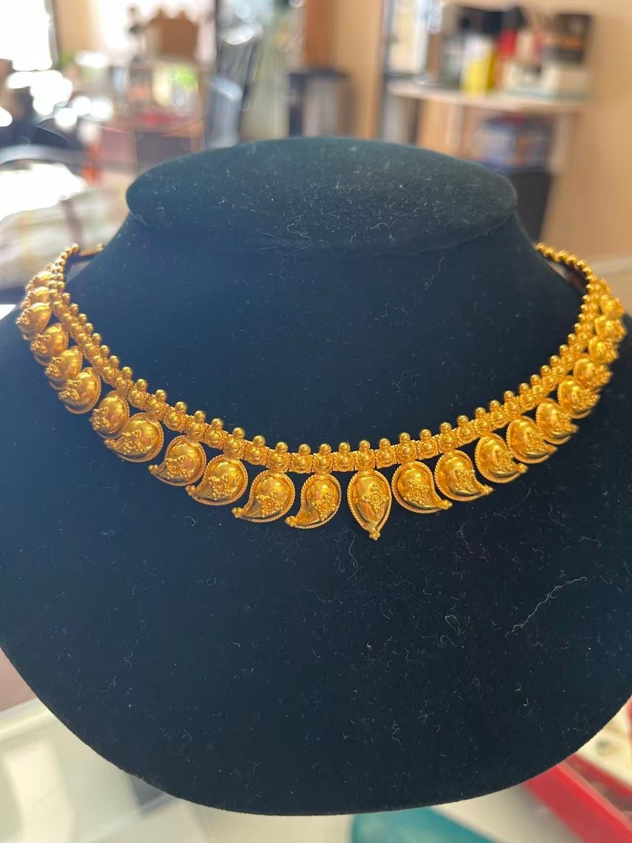 A gold necklace featuring a row of ornate, teardrop-shaped motifs on a black jewelry display bust.