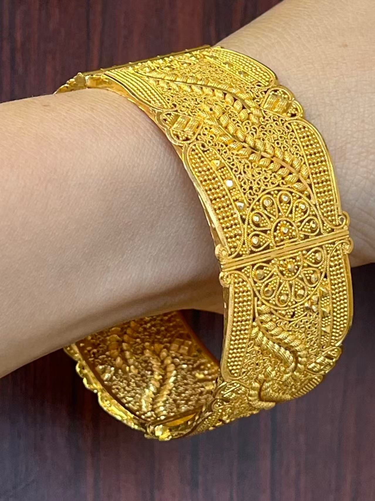 A thick, ornate gold bangle with intricate filigree and floral patterns worn on a wrist against a wooden background.