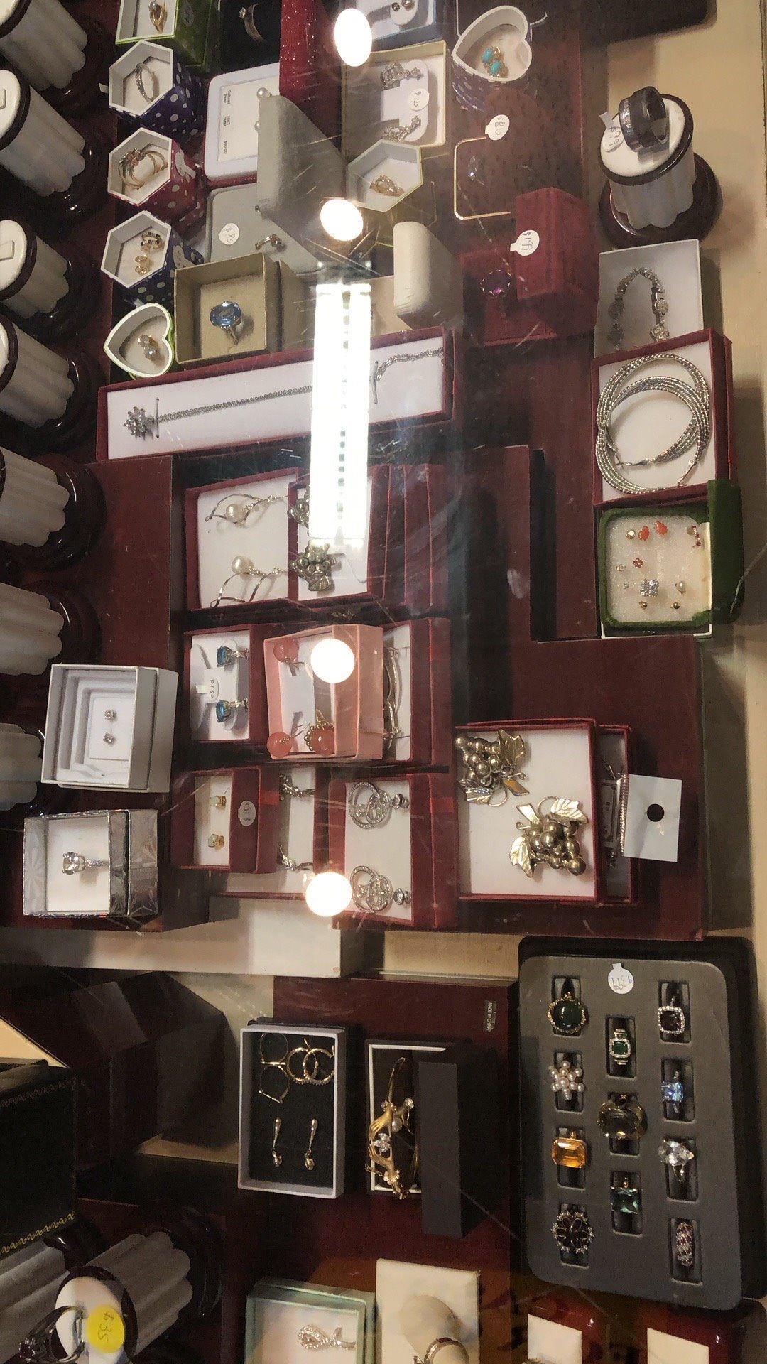 Jewelry display case with various rings, necklaces, bracelets, and earrings. Red-brown background with bright lights.