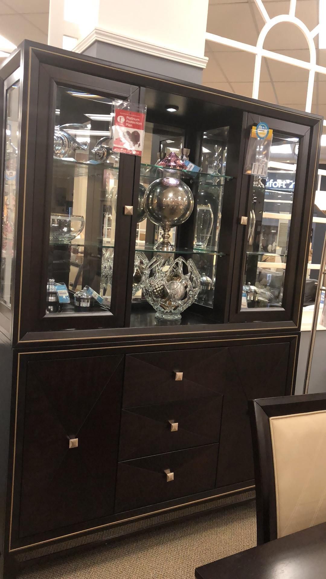 Dark wood display cabinet with glass doors, drawers, and illuminated interior displaying various decorative objects.
