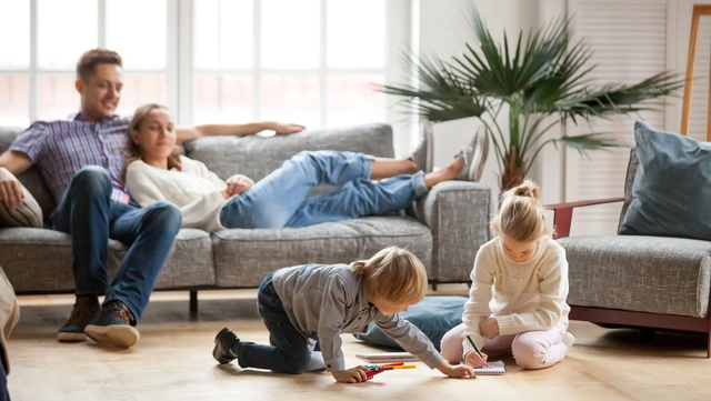 Family relaxing in living room: Dad on couch with daughter, two younger kids playing on the floor.