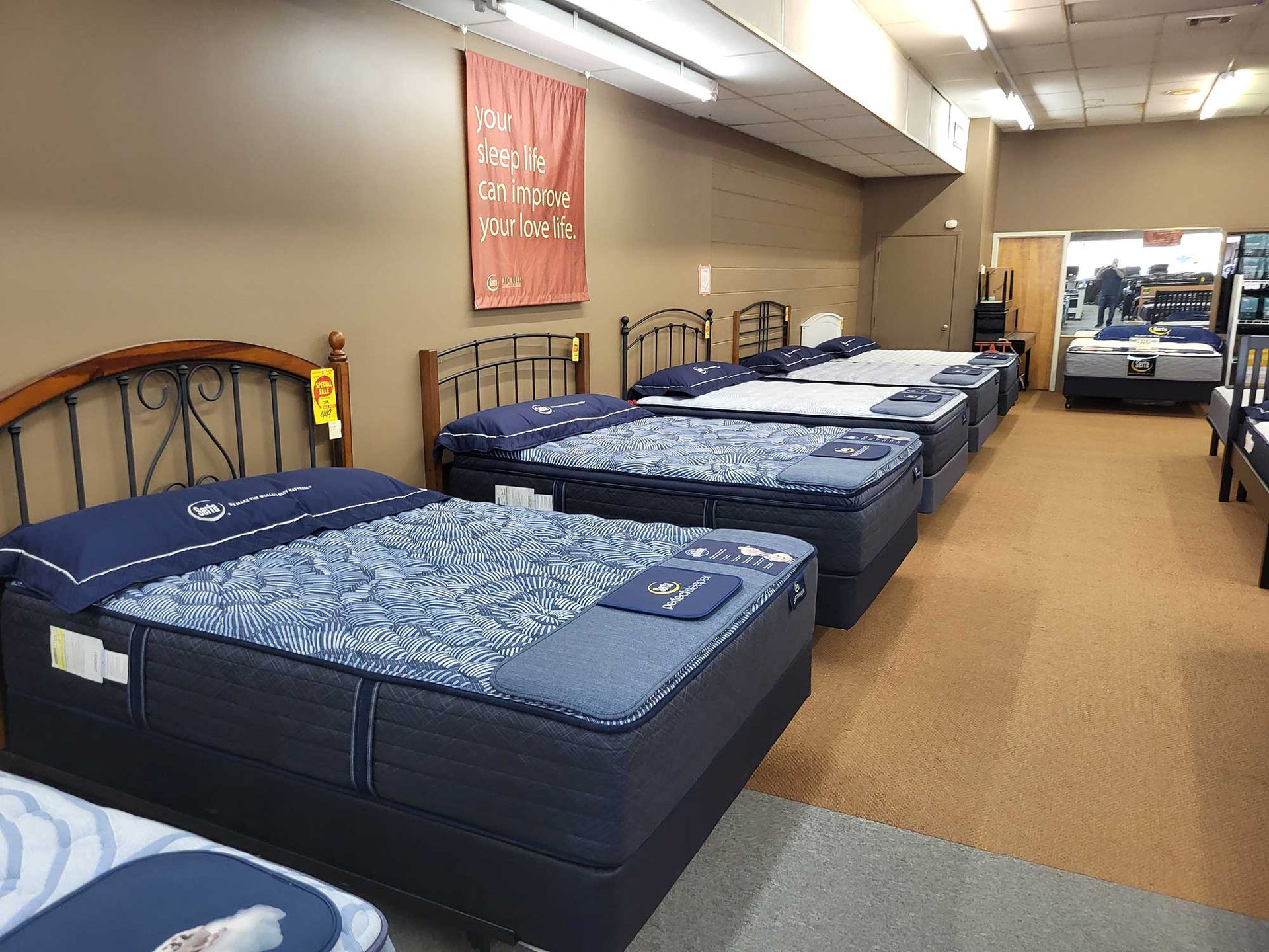 Mattress store interior with several beds on display, beige walls, and brown carpet.