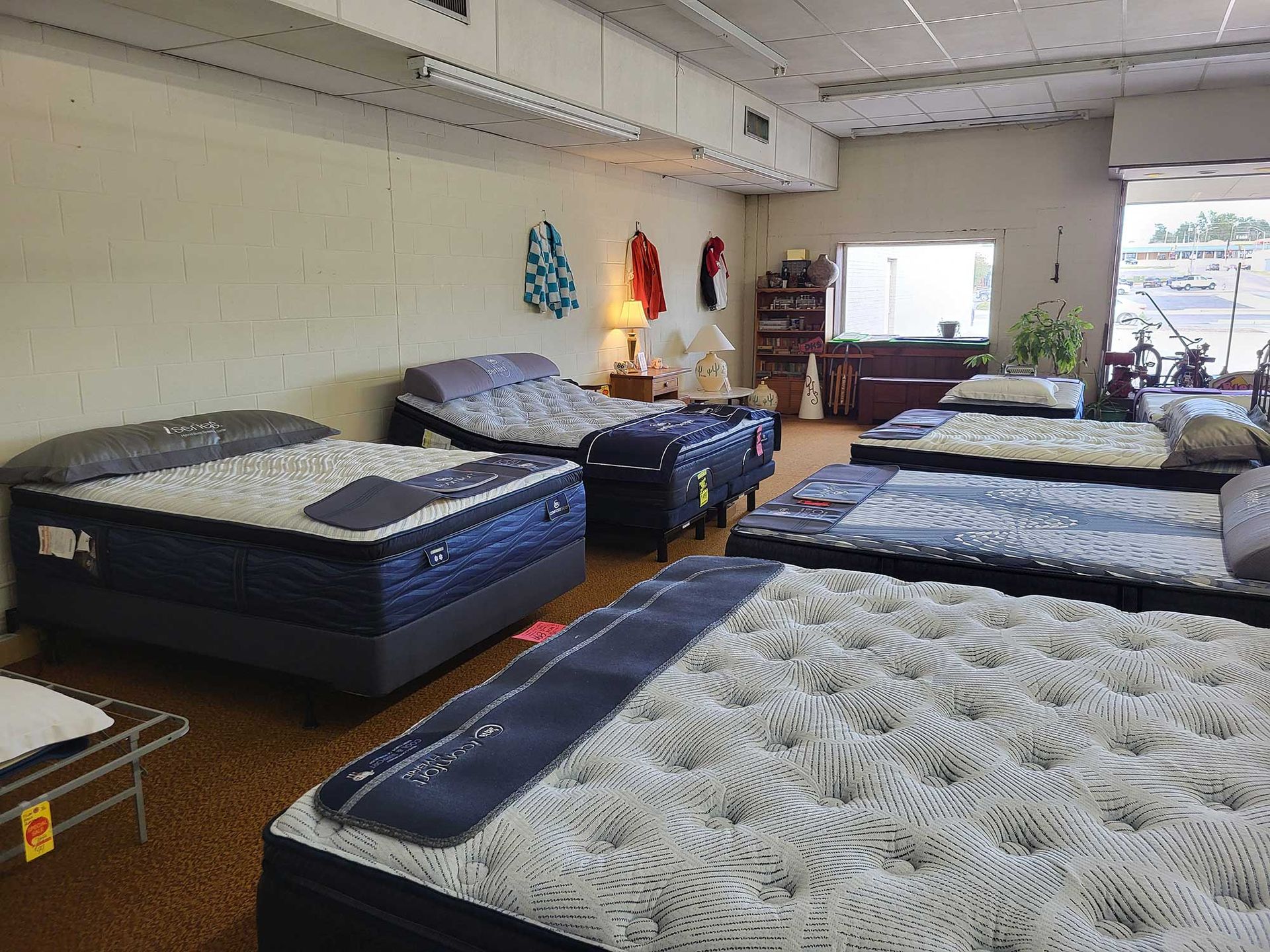 Mattress showroom with multiple beds on display; beige walls, brown carpet, window to the right.