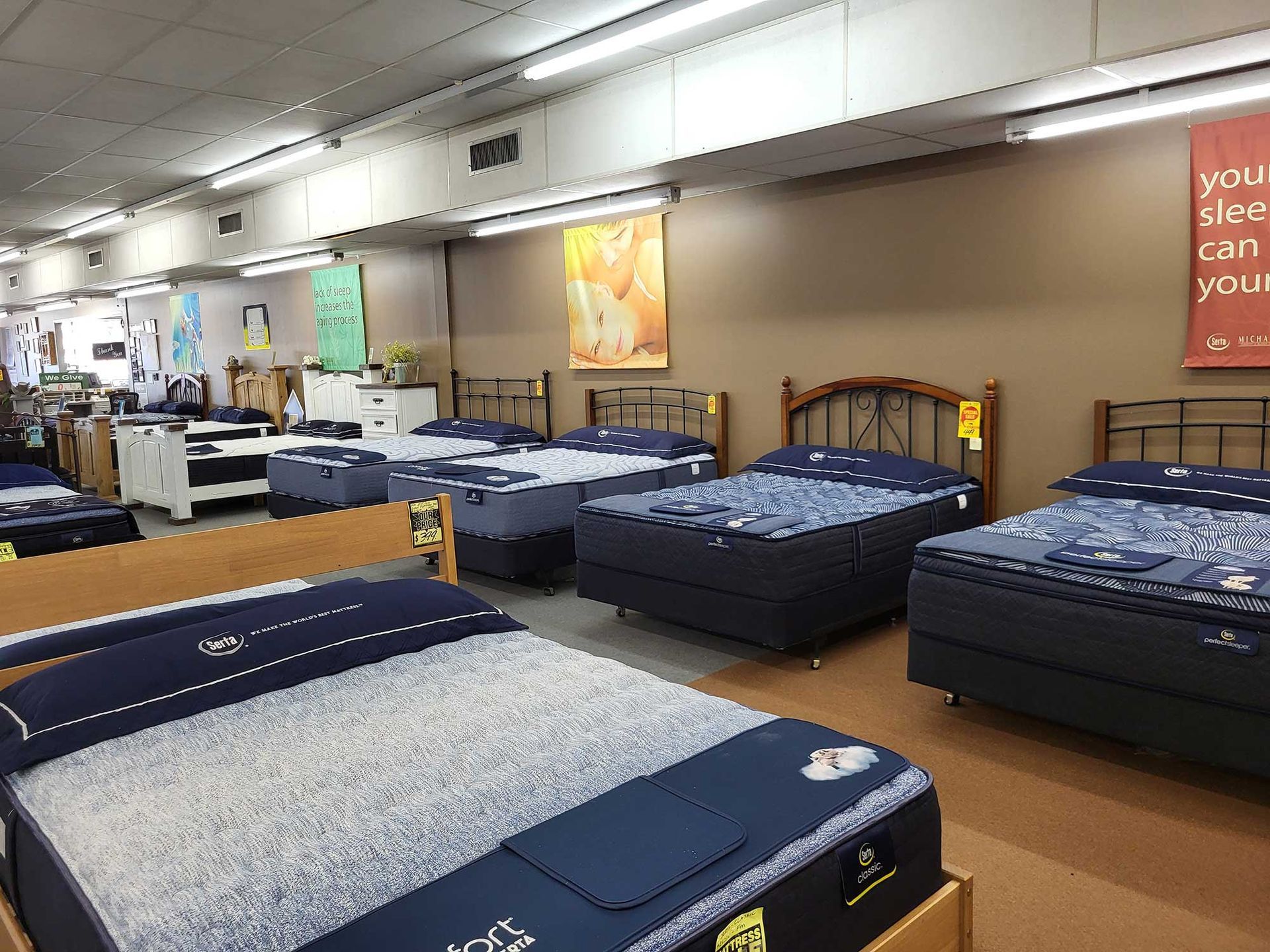 Mattresses on display in a store, with various wooden and metal bed frames, under bright overhead lighting.