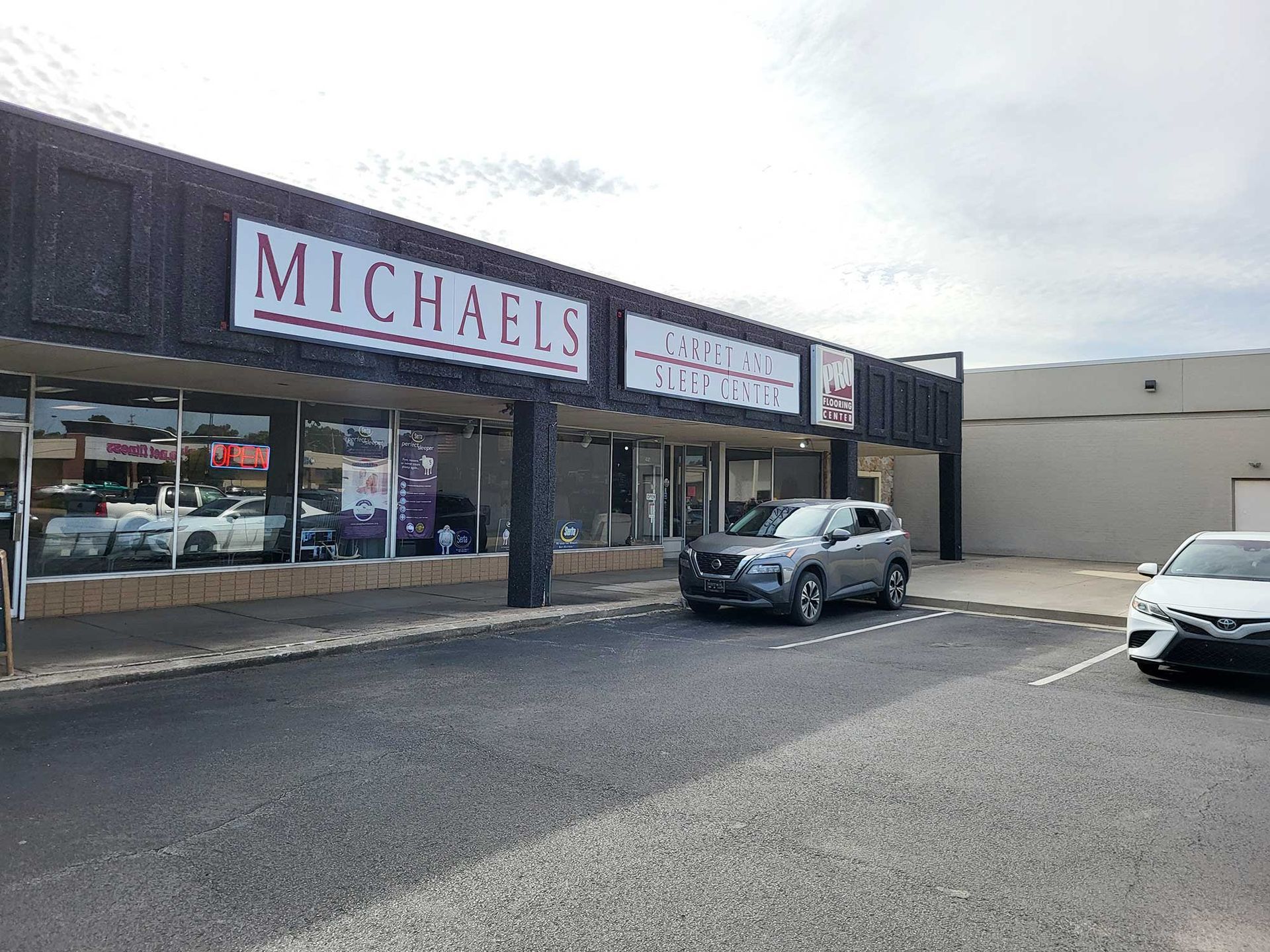 Exterior of Michaels craft store with parked cars. Gray SUV and white car parked in front. Cloudy sky.