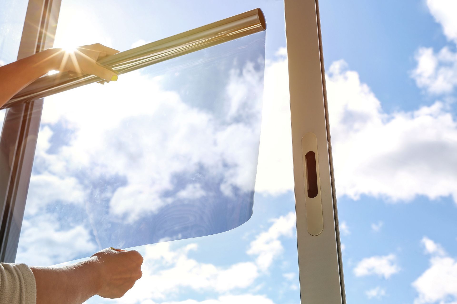 Hands applying a tinted film to a glass window against a bright, sunny blue sky with clouds.