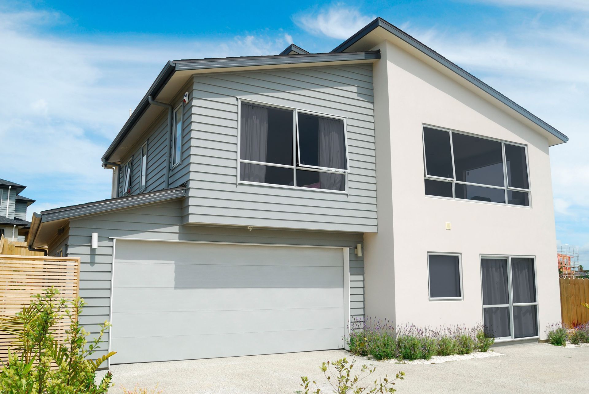 A two-story house with grey horizontal siding, a cream-colored exterior, and a large white garage door under a blue sky.