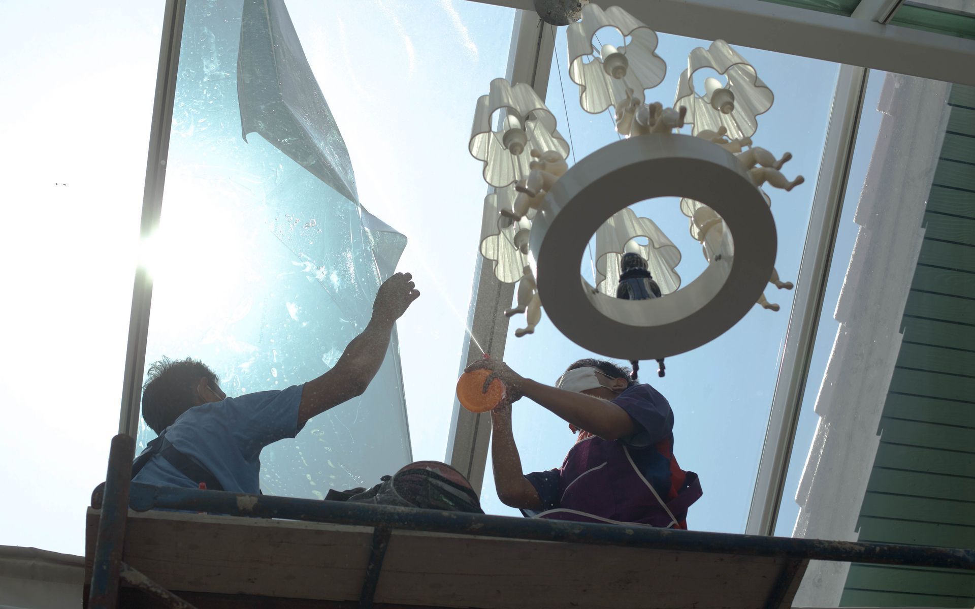Two workers on scaffolding install a white, circular chandelier hanging beneath a glass ceiling in bright sunlight.