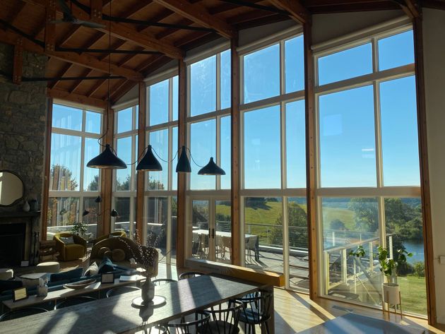 A sunlit room with high ceilings, wood beams, and floor-to-ceiling windows overlooking a green landscape and pond.