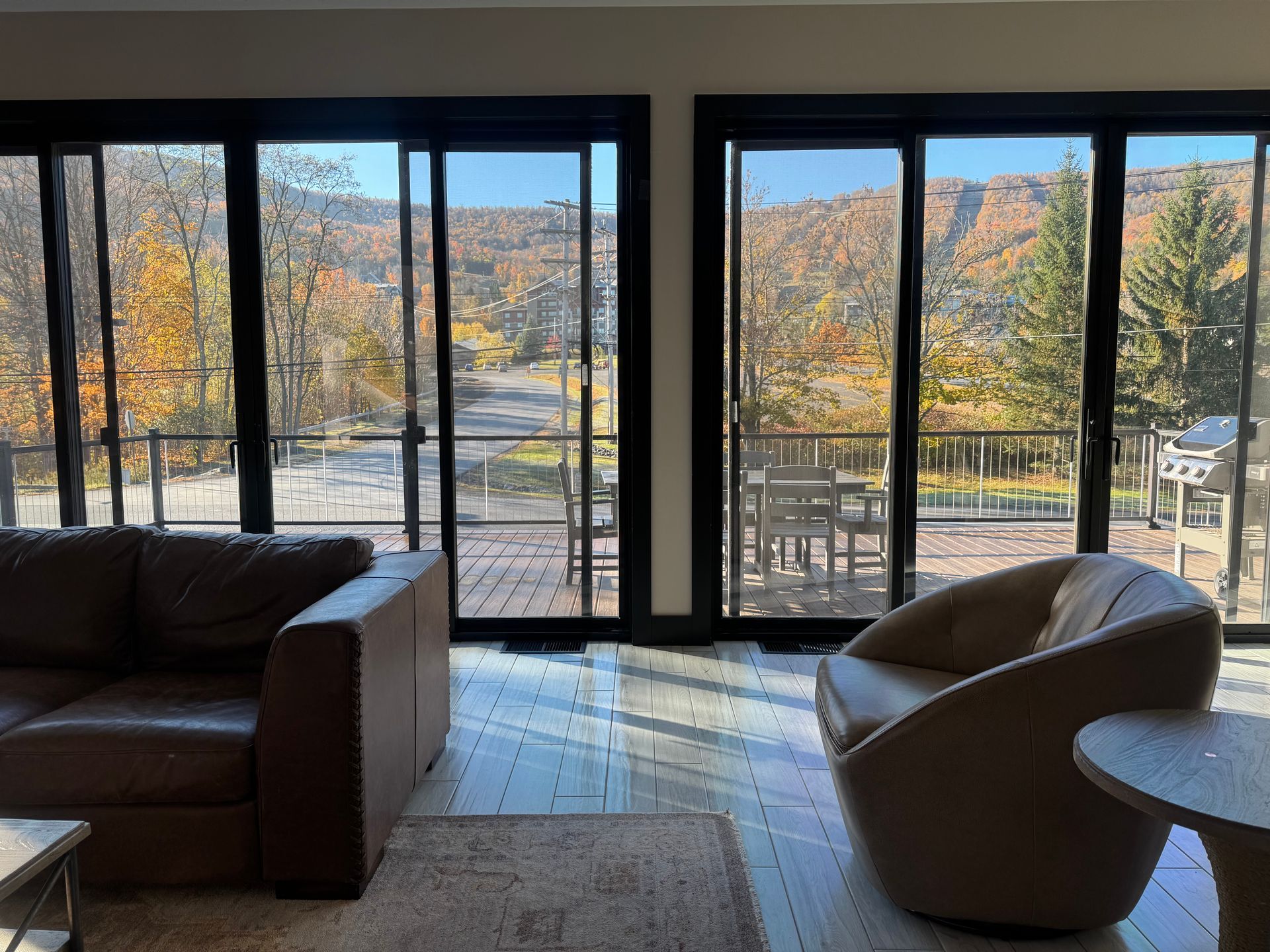 Living room with a brown sofa and armchair facing a balcony with patio furniture and a scenic autumn mountain view.