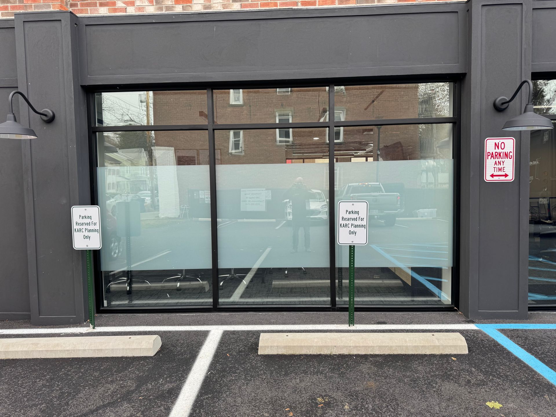 A storefront with dark gray framing and frosted windows, featuring two parking signs in front of striped parking spots.