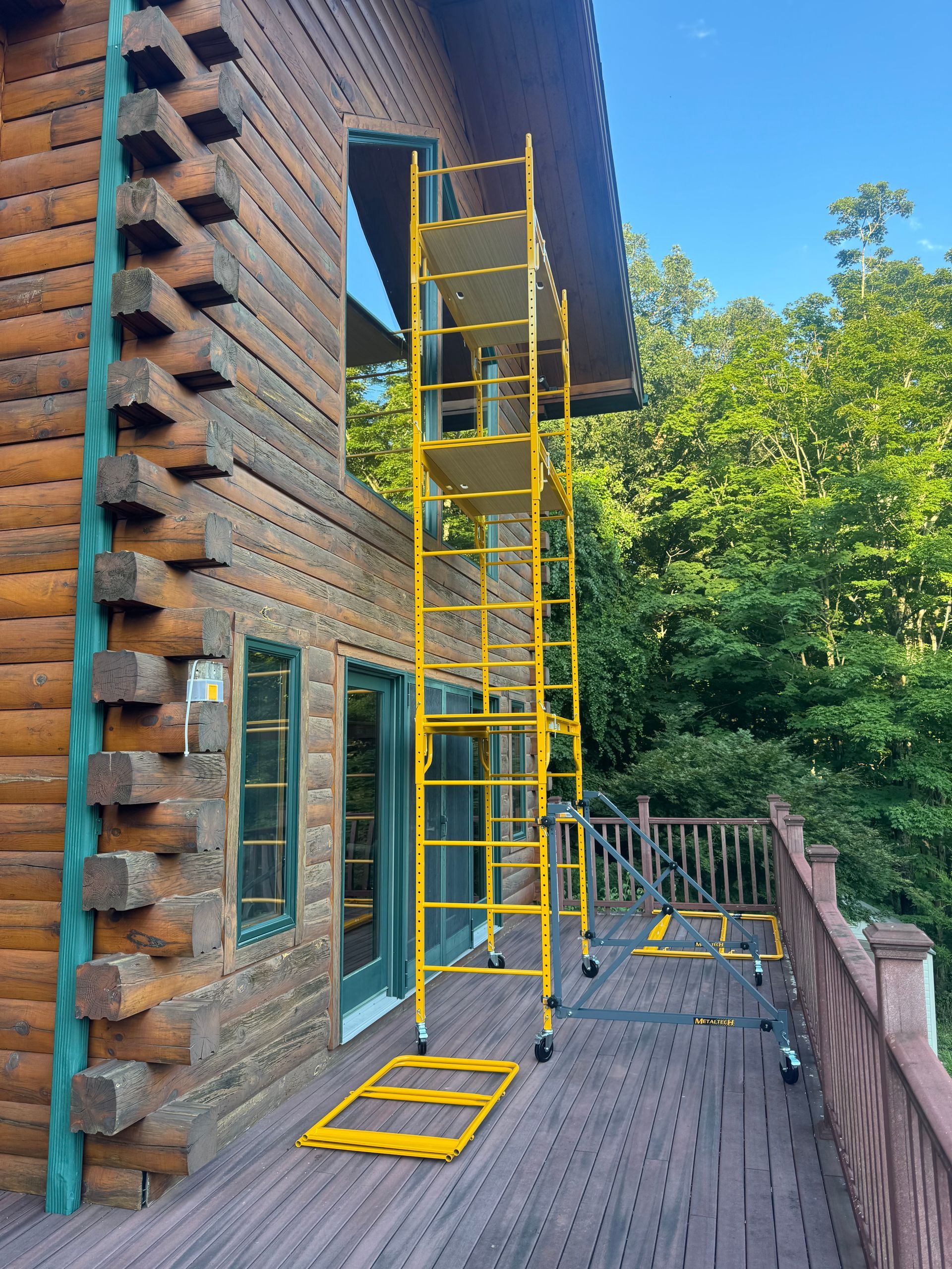 A tall yellow scaffolding unit stands on a wooden deck next to the exterior wall of a log cabin.