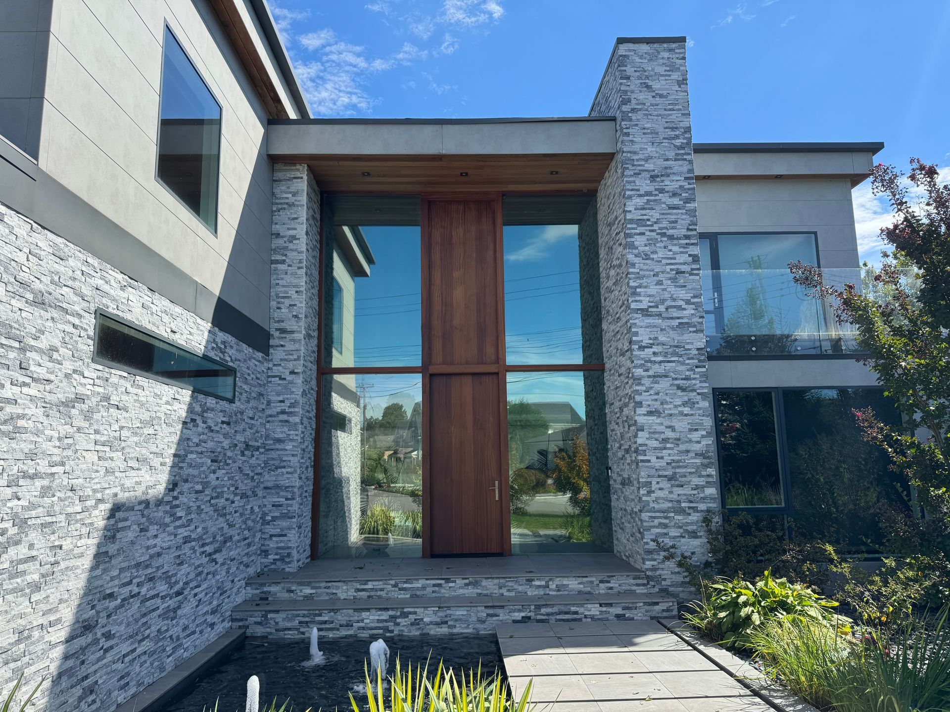 A modern house entrance featuring white stone walls, a tall wooden door, glass panels, and a front water feature.