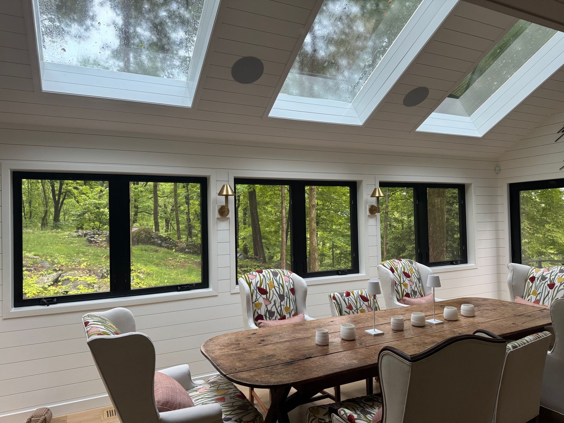A dining room with white-spotted walls, black-framed windows overlooking woods, a wooden table, and upholstered chairs.