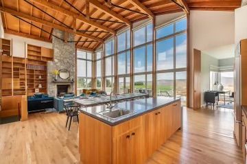 A bright open-plan room with a wood-topped kitchen island, floor-to-ceiling windows, and a stone fireplace.