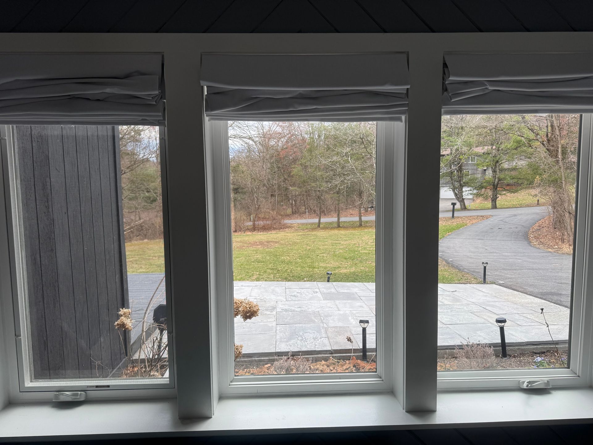 A view through three window panes looking out onto a stone patio, lawn, and driveway surrounded by trees in autumn.