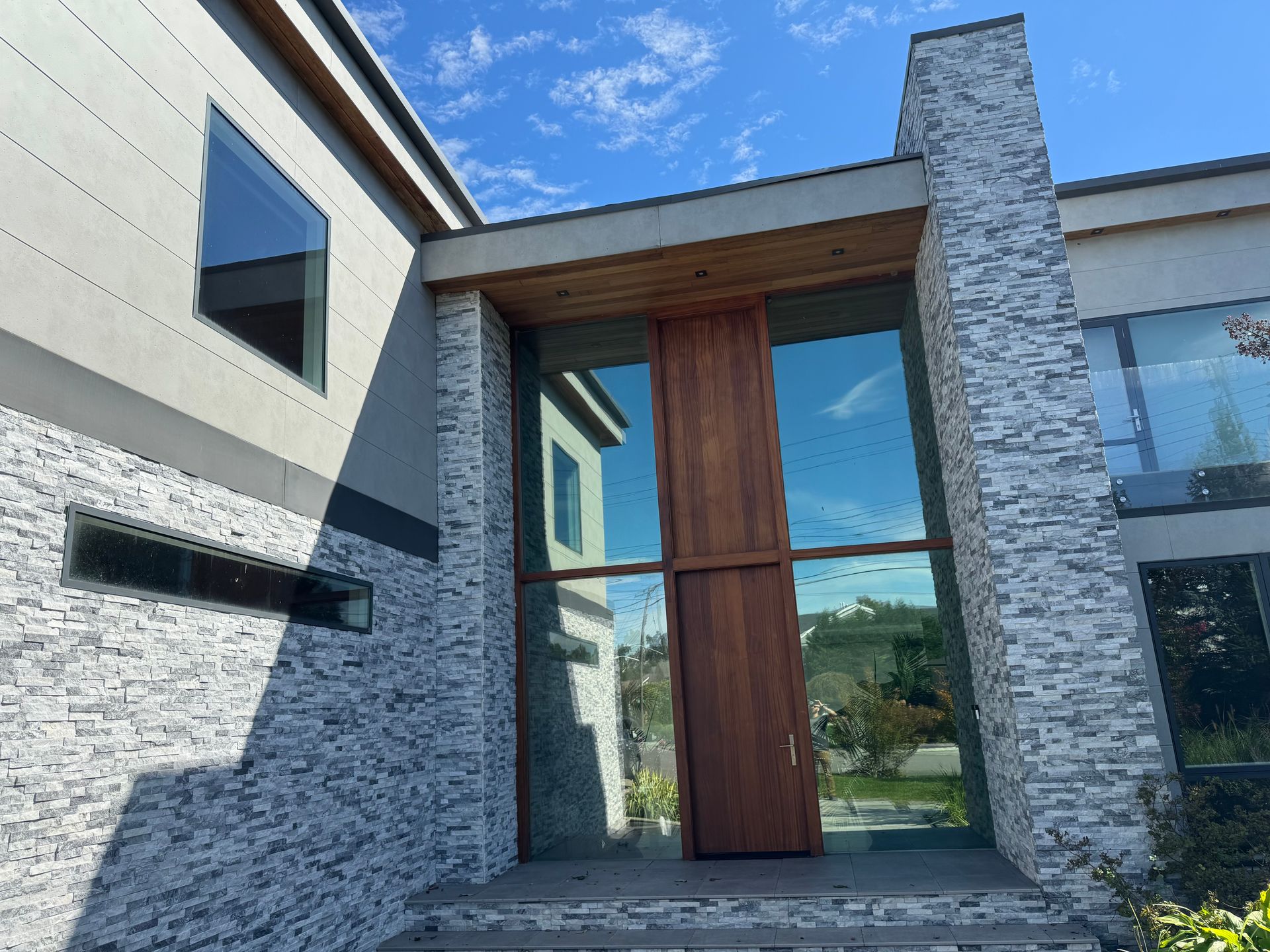 Modern home entrance with a wood door framed by glass, stacked stone walls, and a blue sky in the background.