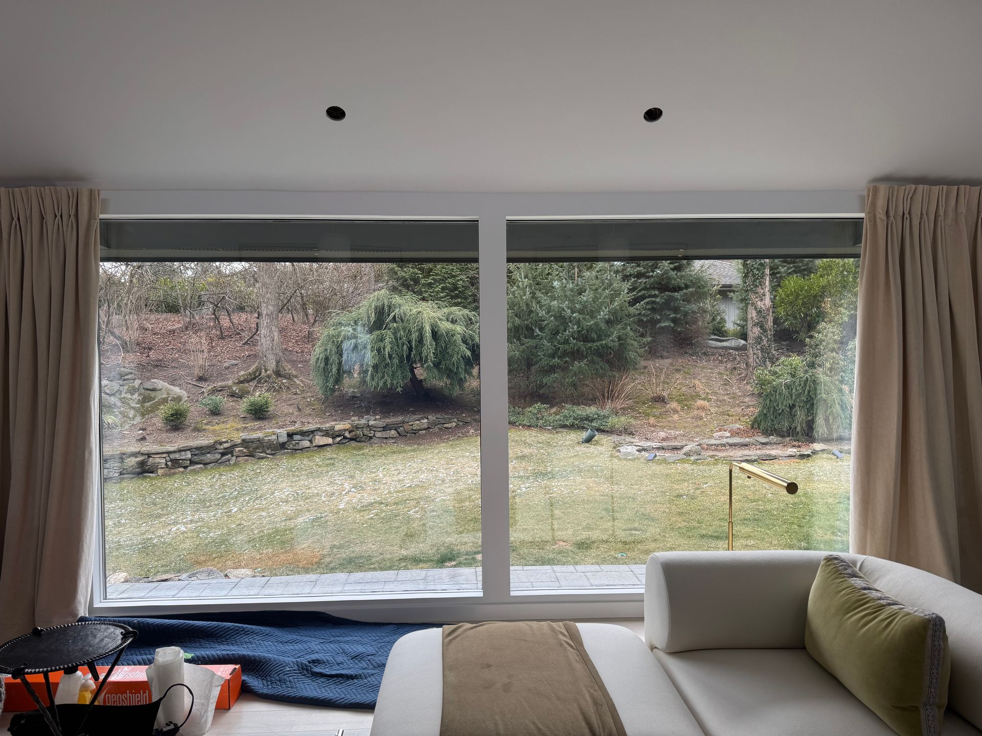 Large picture windows overlooking a grassy backyard with trees, framed by beige curtains and a white couch in the foreground.