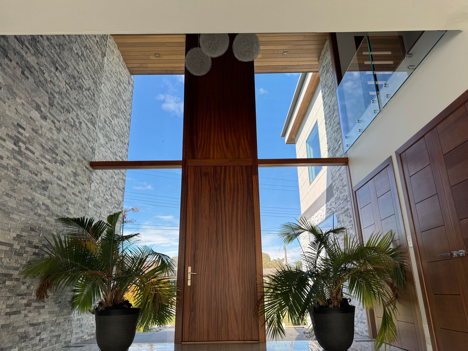A tall, wooden front door framed by two large potted plants, flanked by a stone wall and a modern glass-paneled staircase.