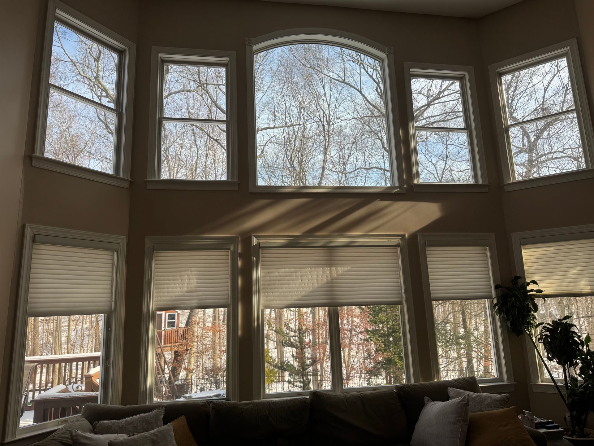Two rows of windows in a living room look out onto a snowy, wooded yard; the bottom row has partially closed shades.