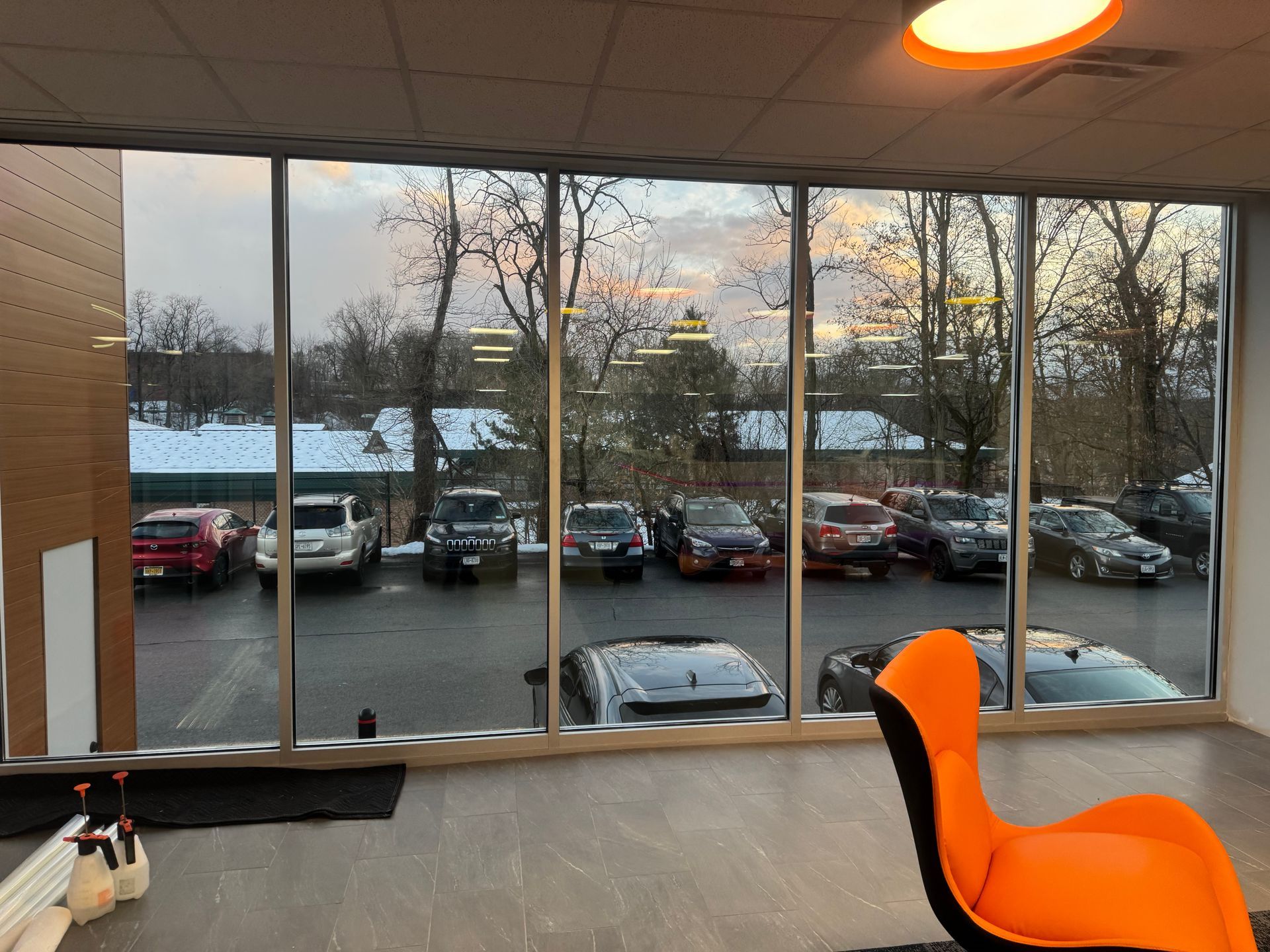 A bright orange chair sits in an office space facing a floor-to-ceiling window overlooking a snowy parking lot with cars.