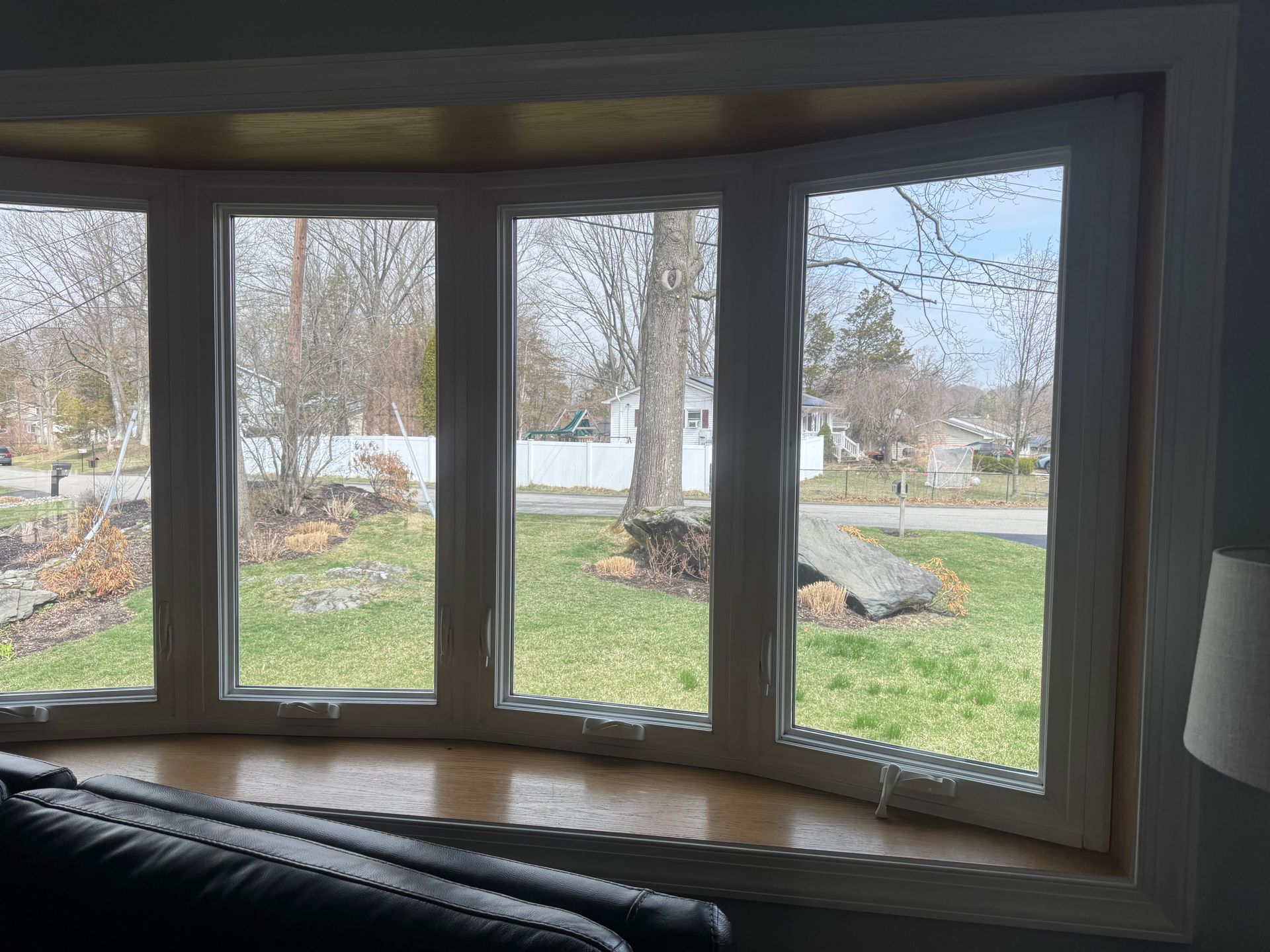 A large, four-paneled bow window looks out over a residential lawn with a white fence, bare trees, and a cloudy sky.