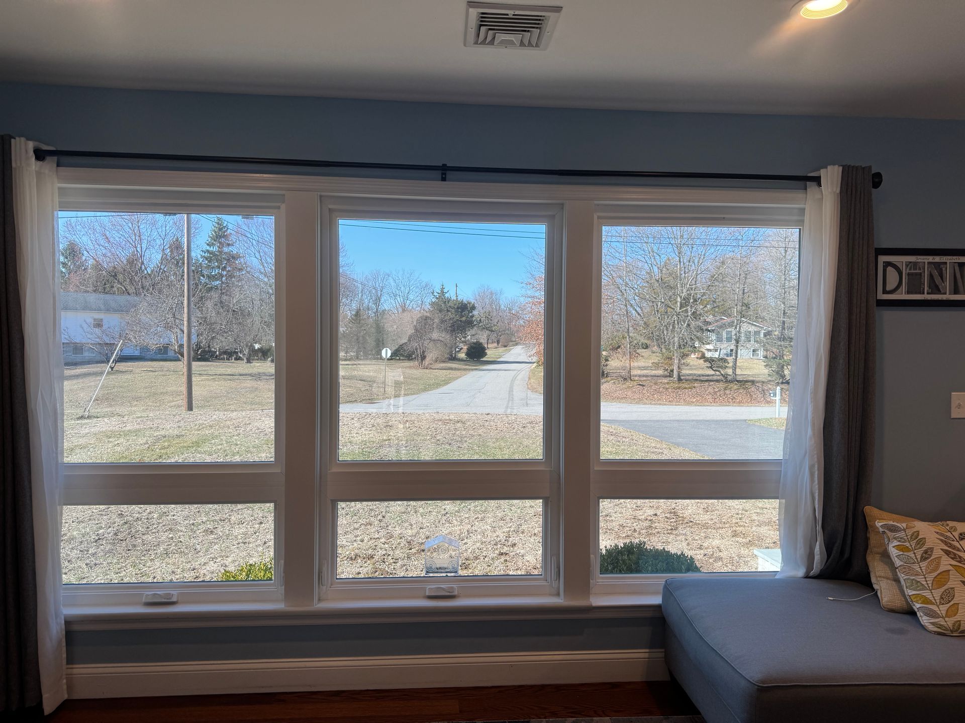 A large, triple-pane window with white frames looks out onto a suburban yard, framed by dark grey and white curtains.