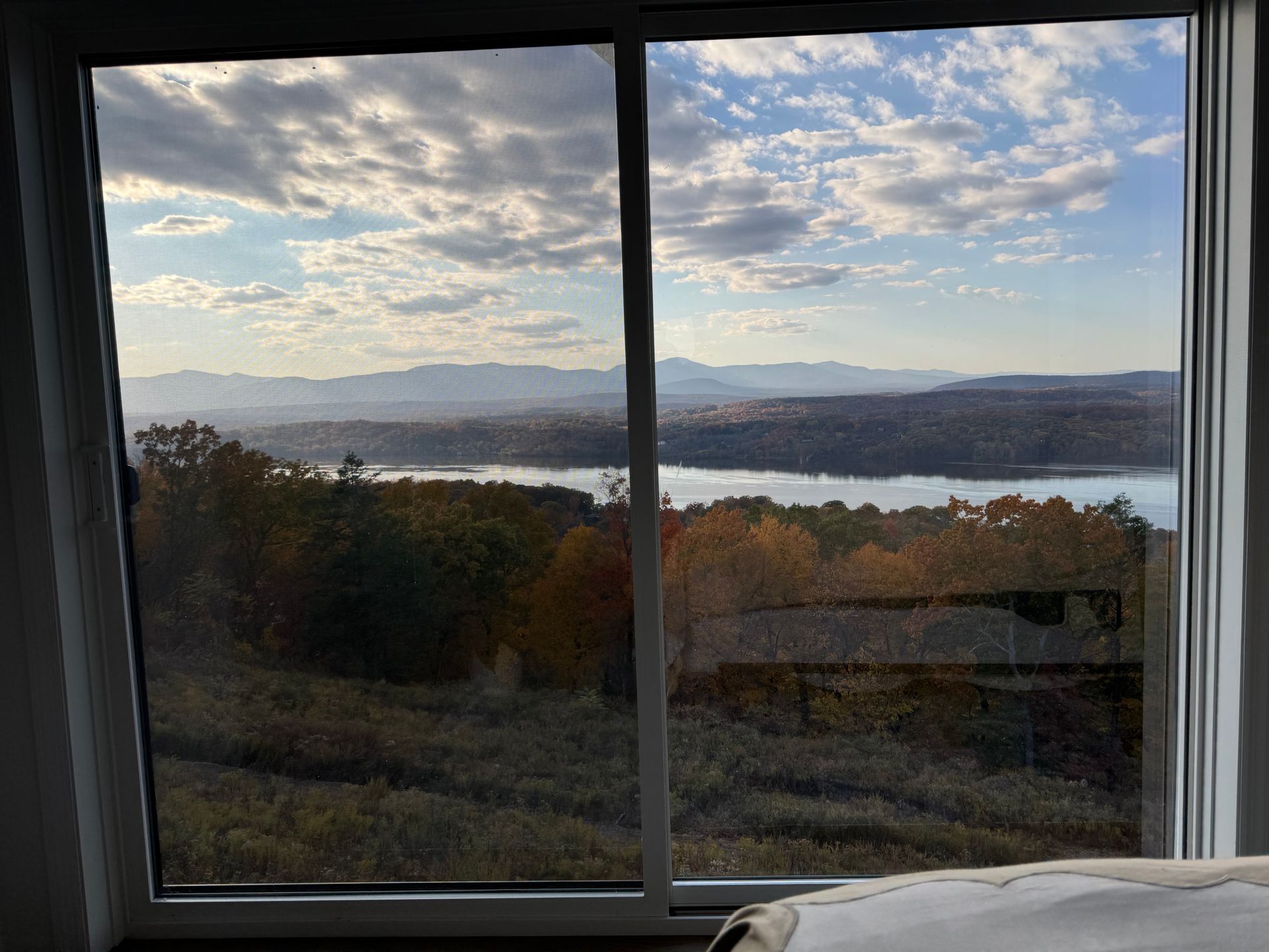 A window view of a river and distant mountains framed by autumnal trees under a cloudy blue sky.