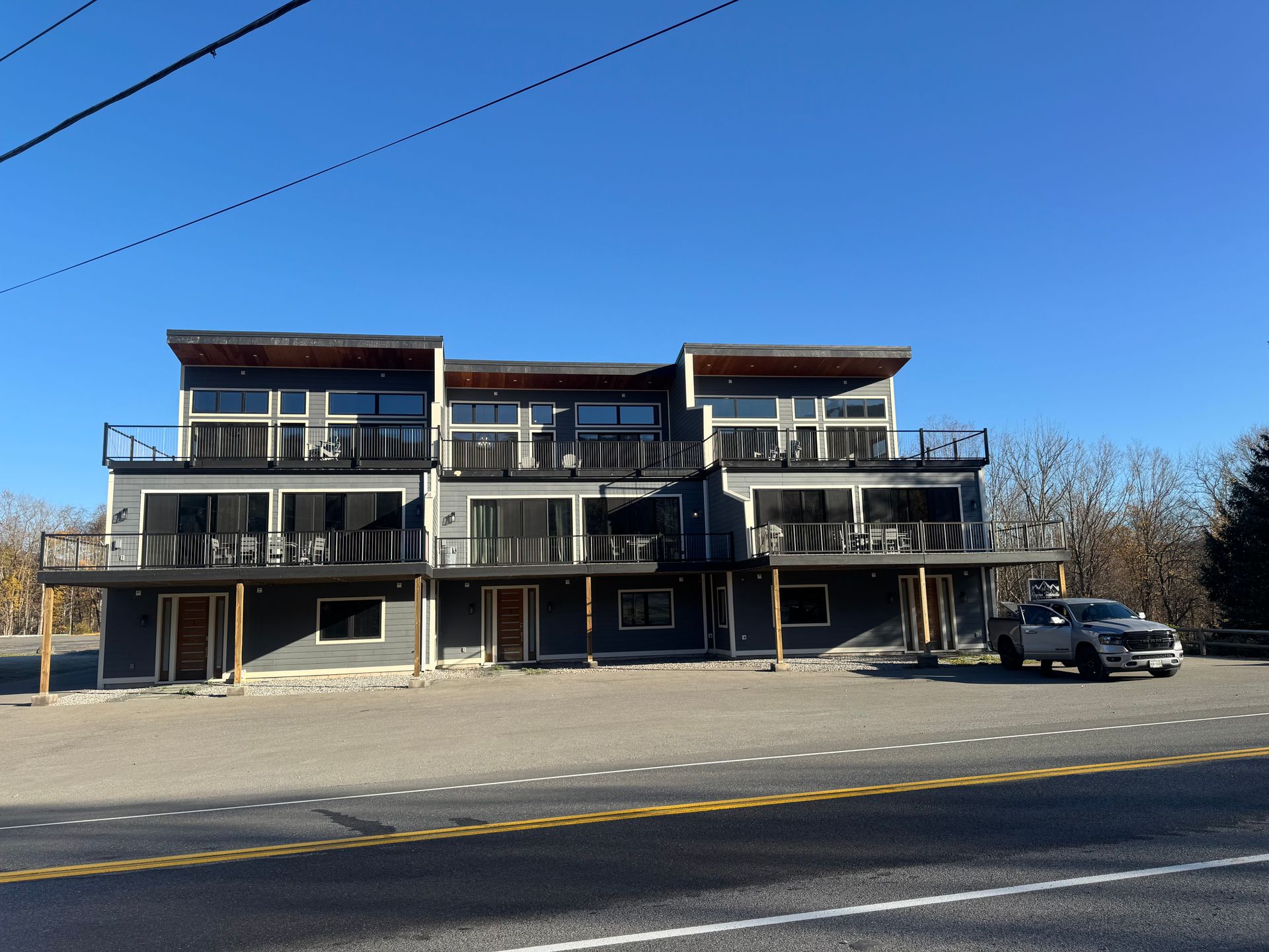 A modern, multi-story gray building with wooden balconies and large windows faces a parking lot and a road.