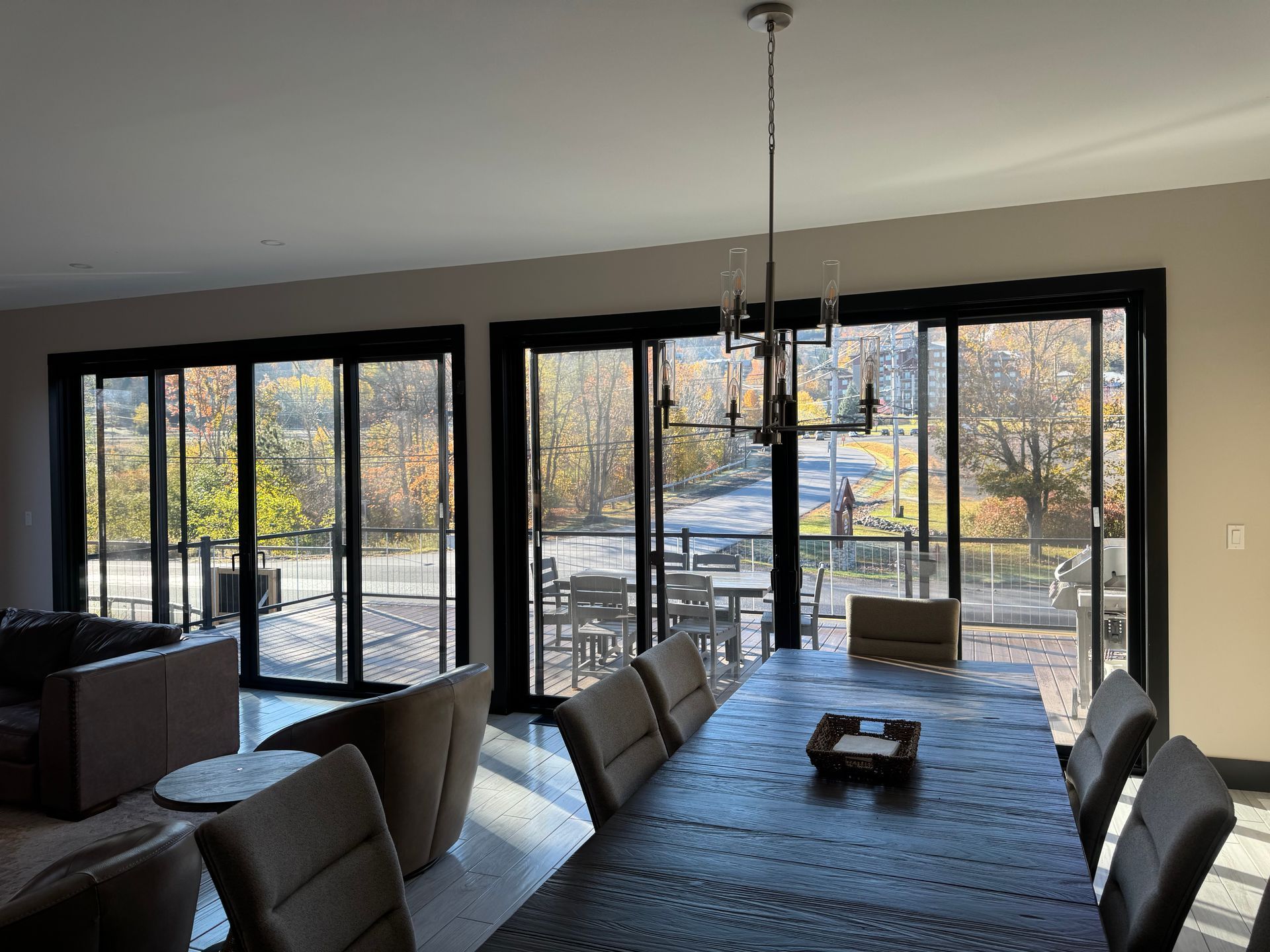 A dining room with a long dark wooden table and chairs, facing floor-to-ceiling glass sliding doors looking onto a deck.