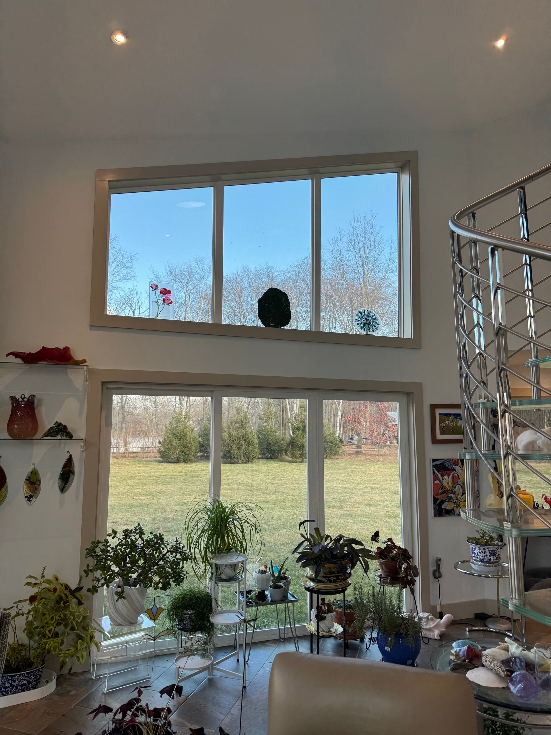 Large, stacked windows look out over a sunny field, surrounded by potted plants and a spiral staircase in a home interior.
