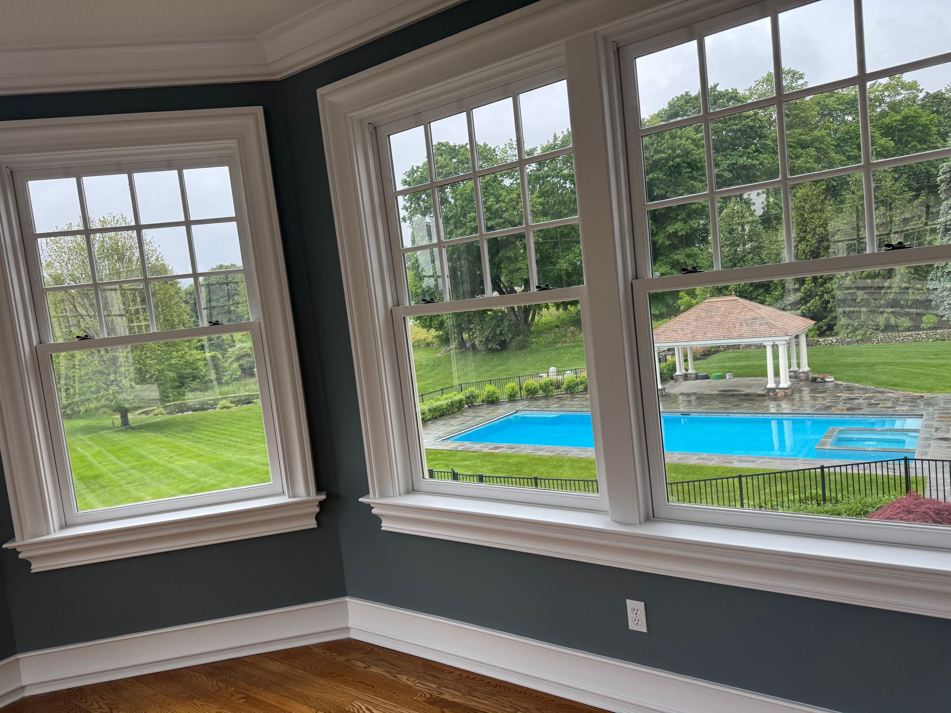 A view from a room with dark gray walls and white trim, looking out through large windows at a backyard pool and gazebo.
