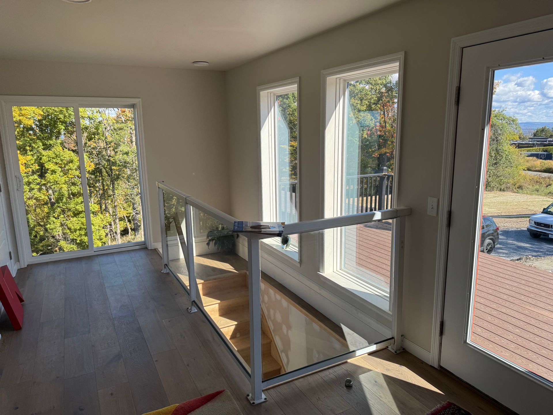 An interior view of a room with hardwood floors, a white staircase railing, and large windows looking out to trees.