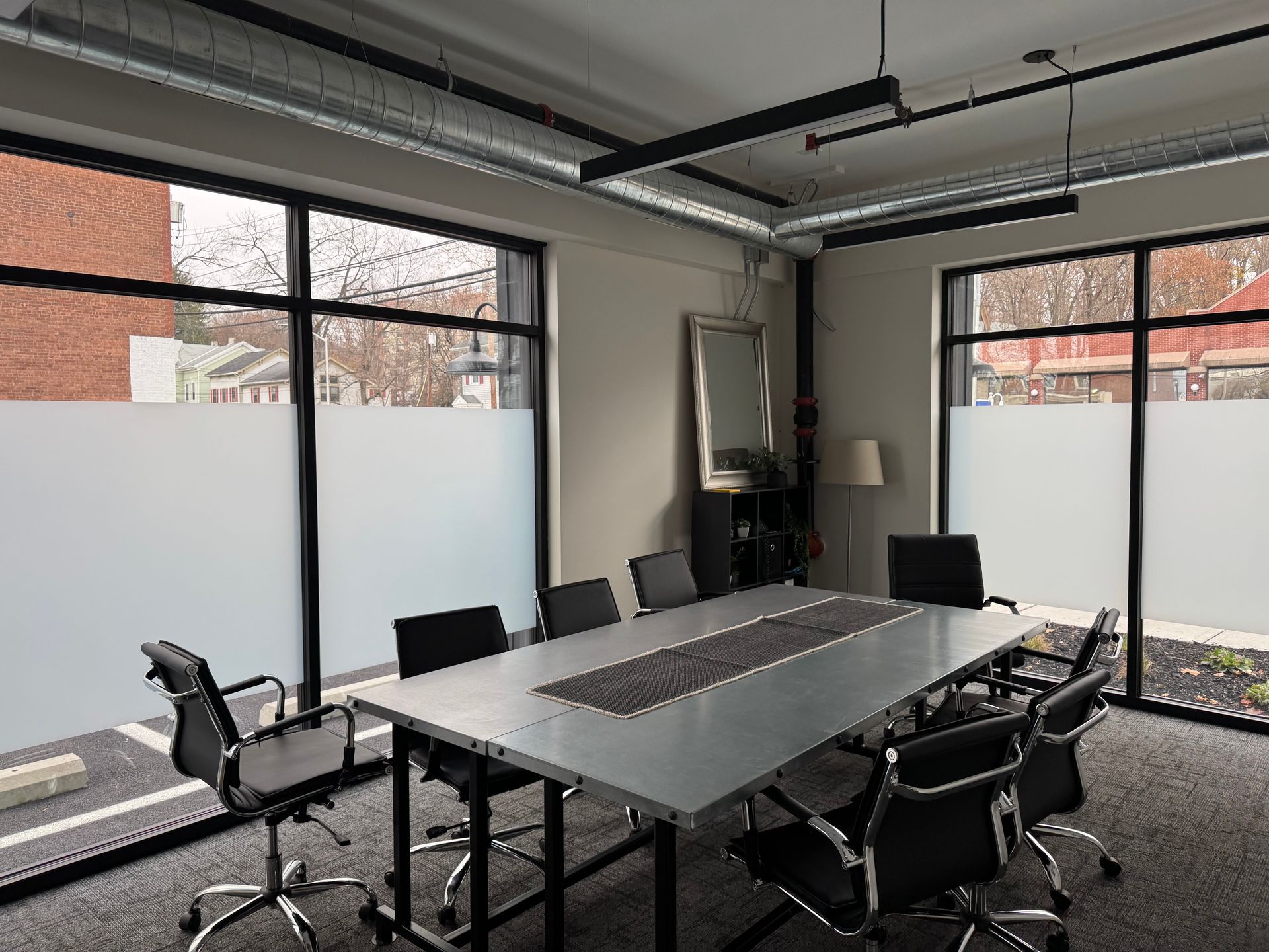 A modern conference room with a long gray table, black rolling chairs, and large windows with frosted privacy film.