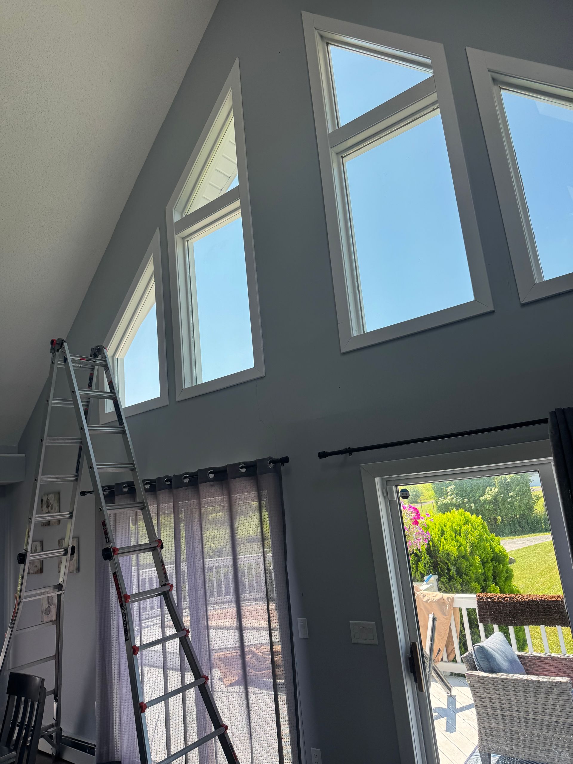 An extension ladder leans against a high interior wall with tall, slanted windows and a view of a deck and trees outside.