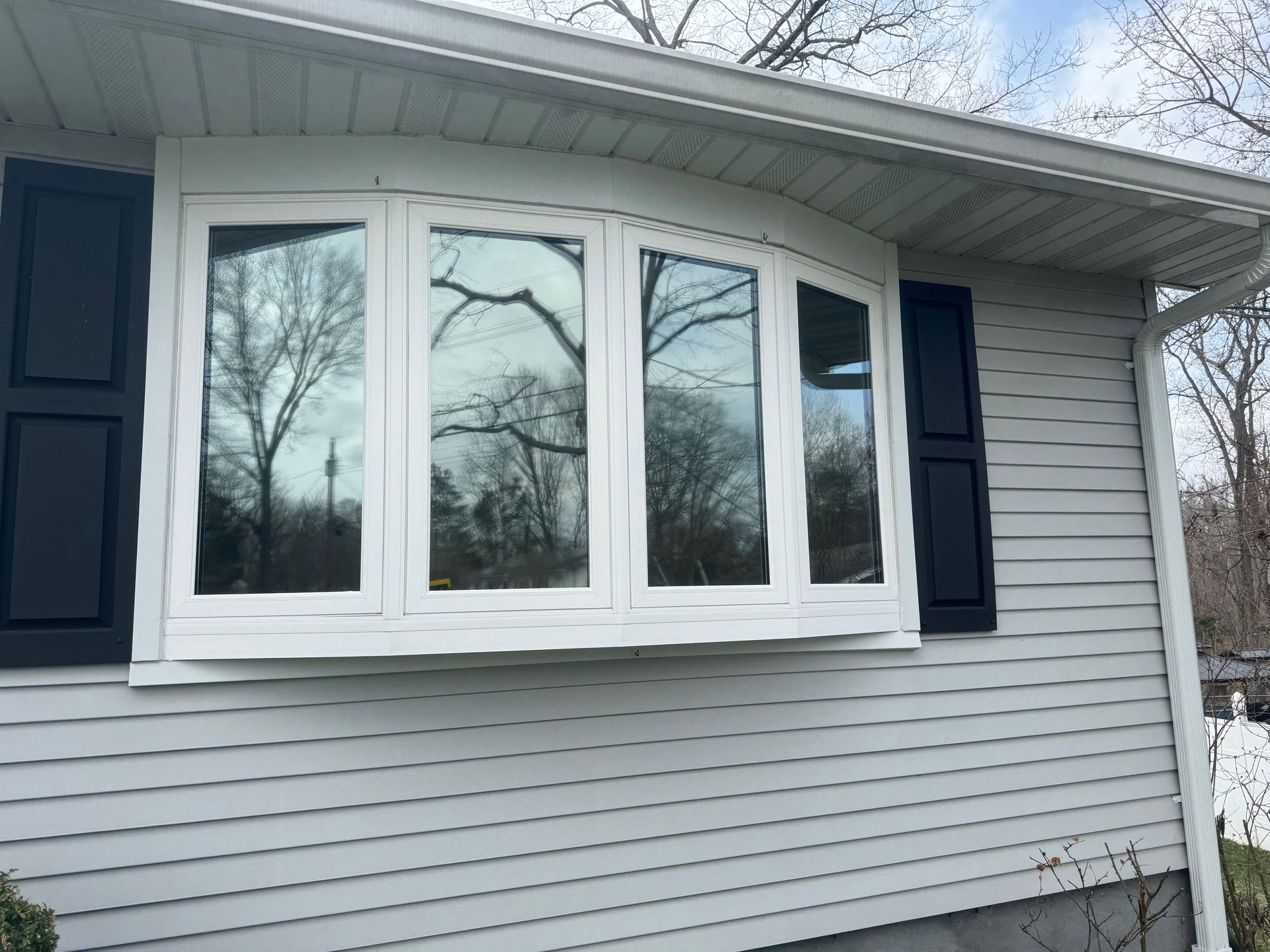 A white bay window with black shutters installed on a light gray vinyl-sided house exterior under a cloudy sky.