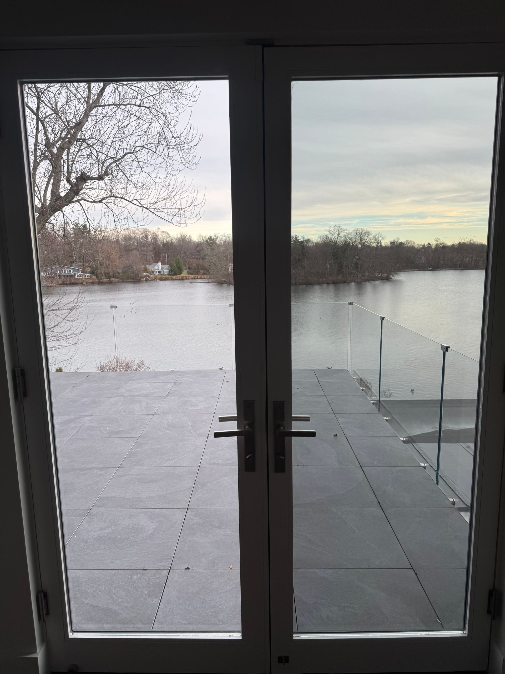 A view through double glass doors looking out onto a dark patio deck, a glass railing, and a lake at dusk.