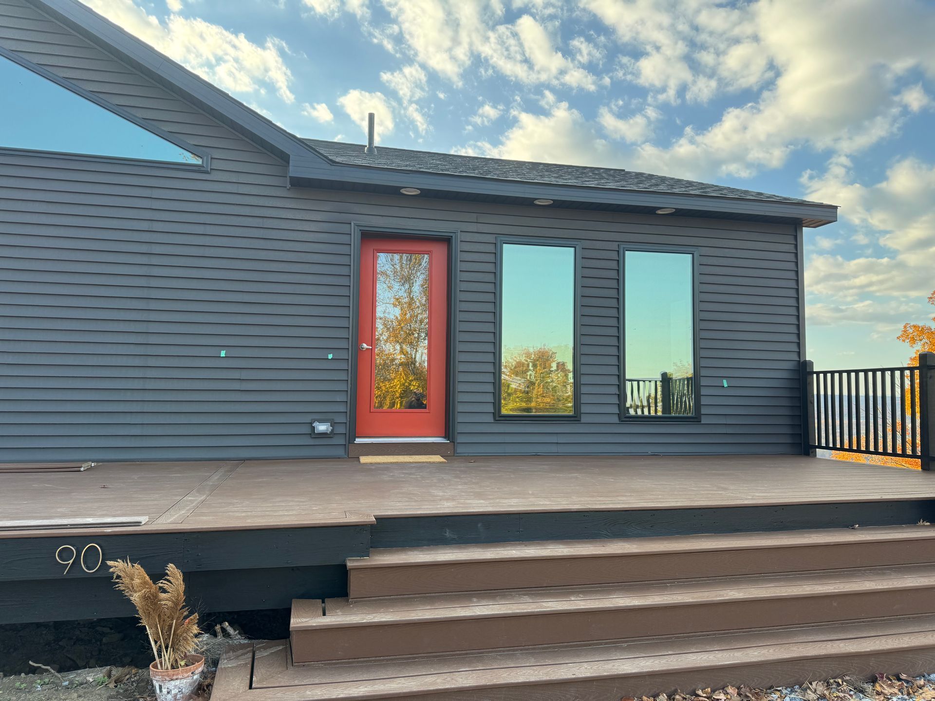 A dark-sided modern house with a bright red front door, two large windows, and wooden stairs leading to a deck.