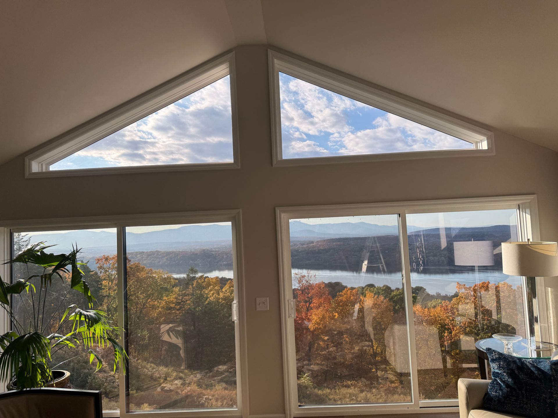 Large windows with triangular top panels frame a scenic view of a body of water and trees with autumn foliage.