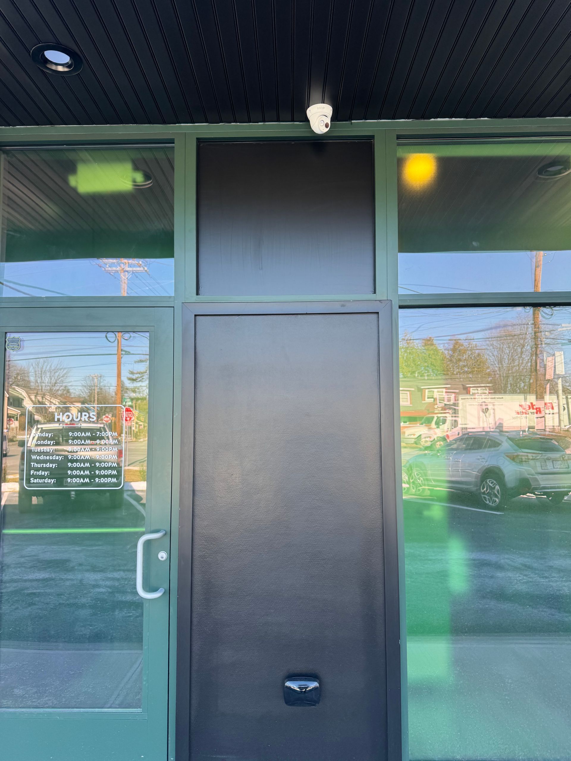A commercial entrance with a dark door, a glass door with business hours, and large windows under an outdoor light.