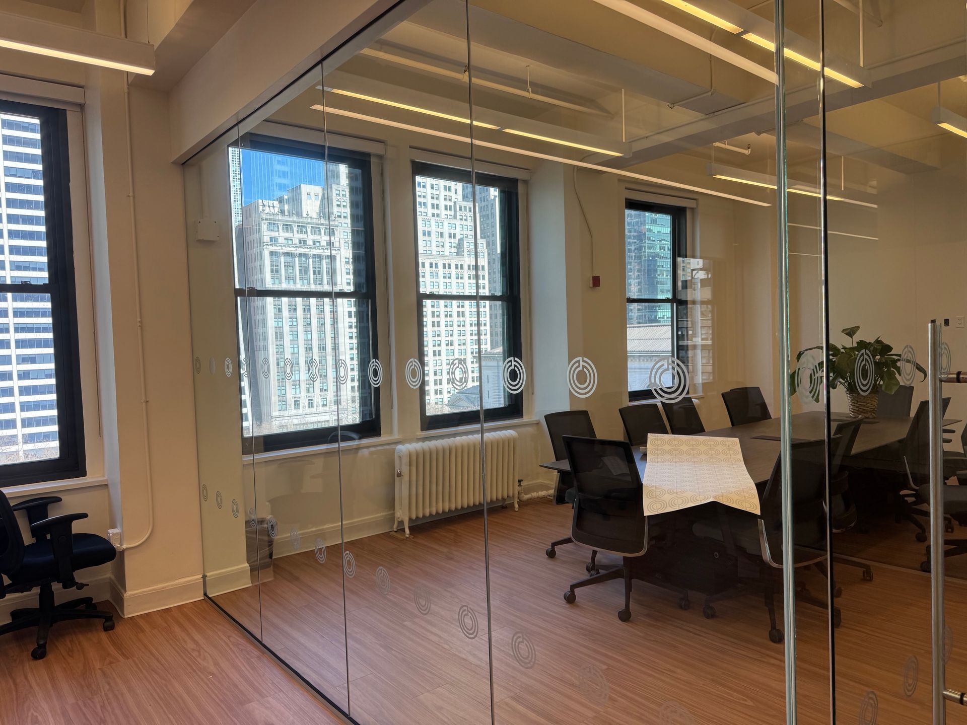 An office meeting room with a wooden table, ergonomic chairs, glass walls, and large windows overlooking city buildings.