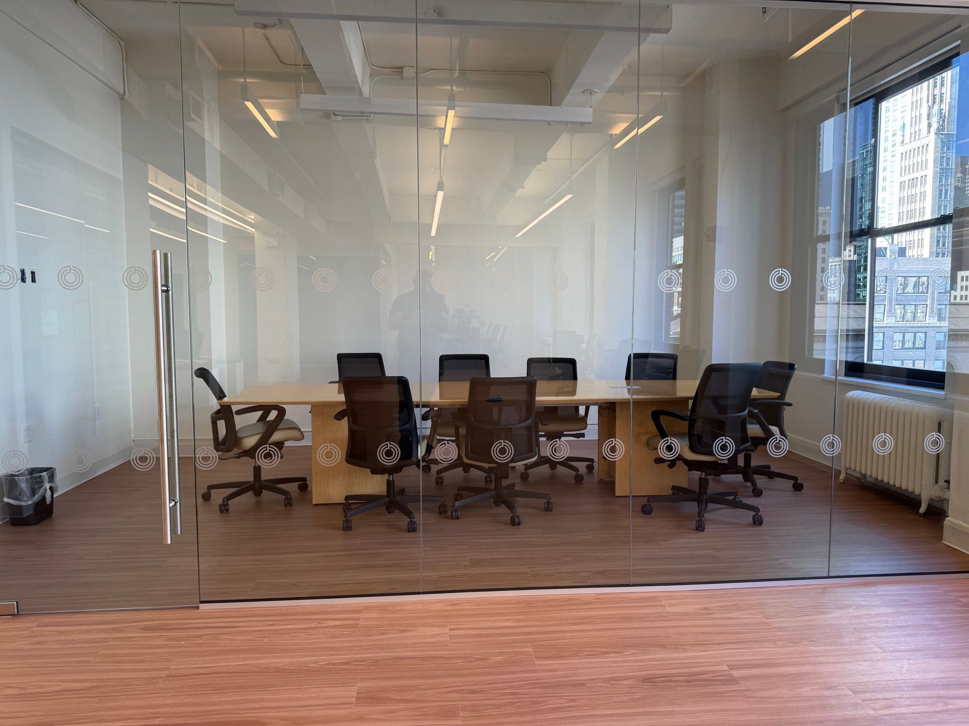 A glass-walled conference room with a wooden table, eight black chairs, and a window view of an urban building.
