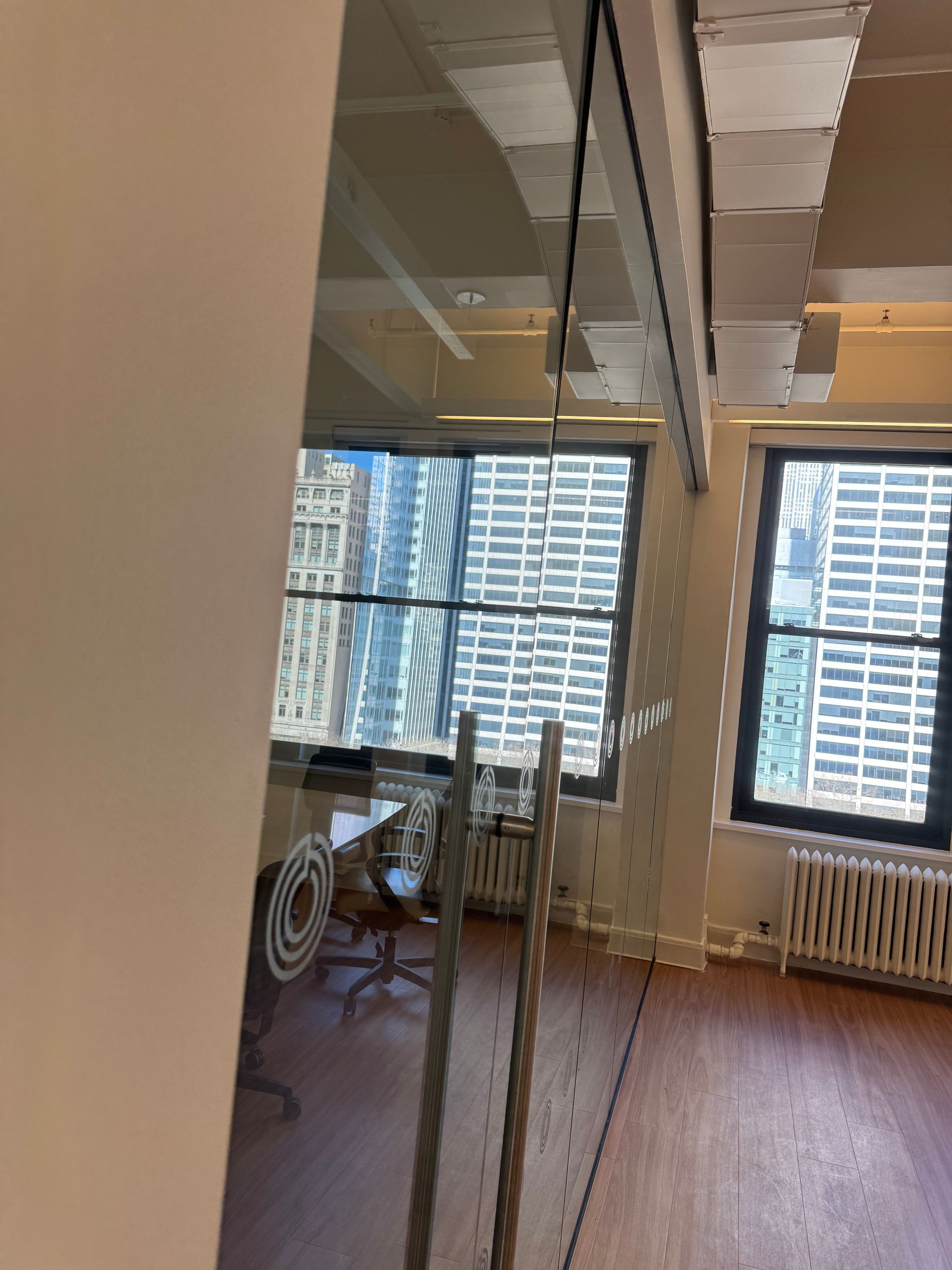 An office interior viewed through glass doors, featuring wooden floors, desks, chairs, and large windows with city views.