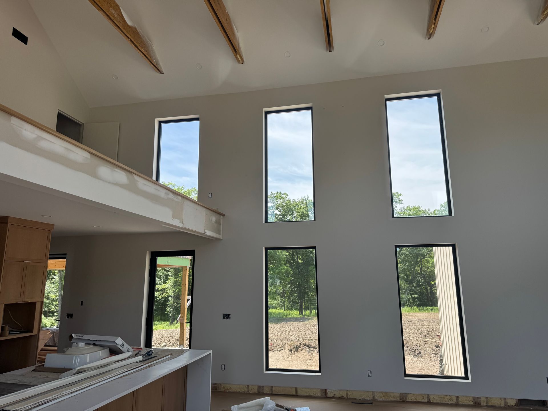 A bright, unfinished room with high ceilings, exposed wood beams, a partial loft, and large, tall rectangular windows.