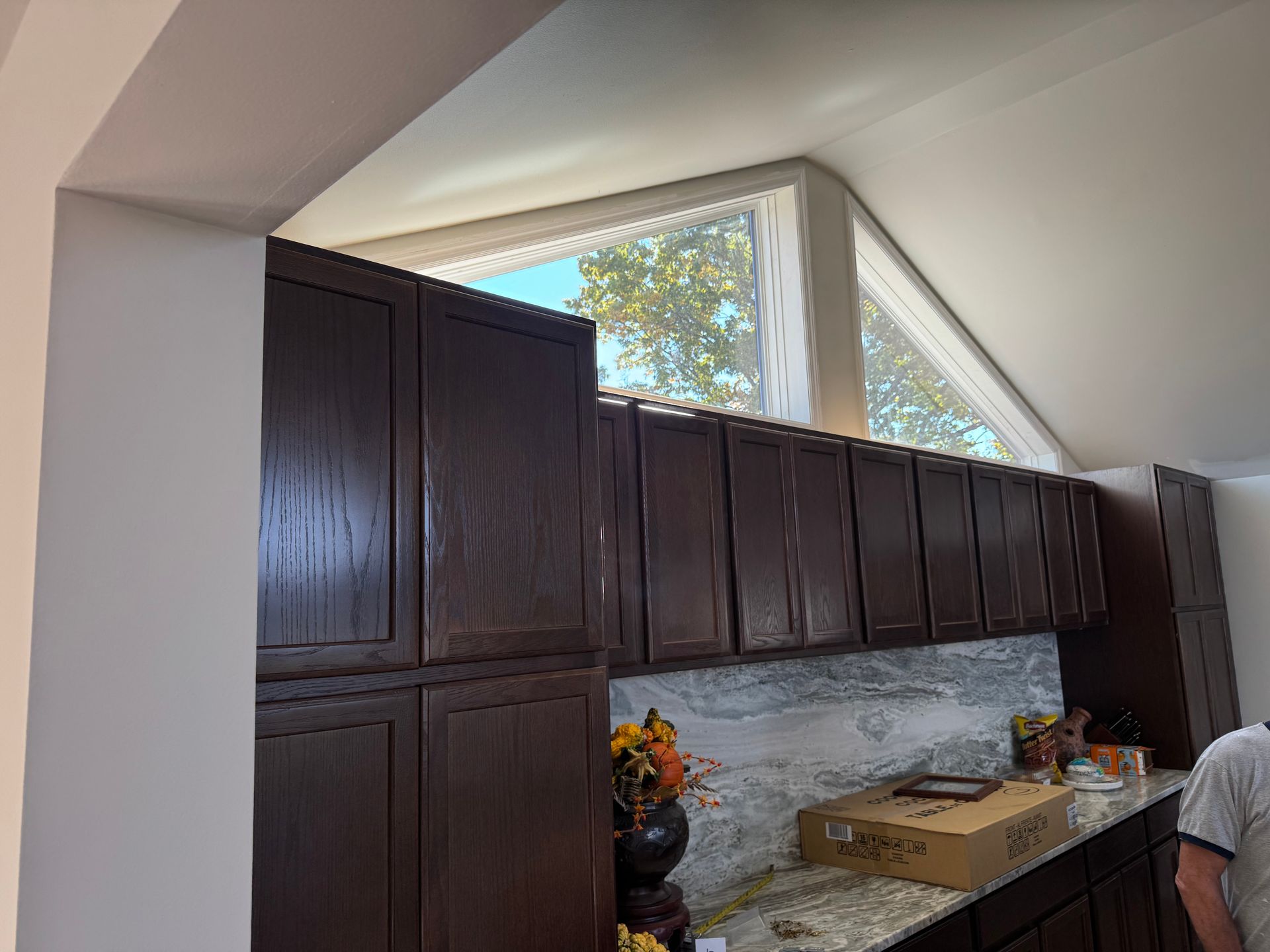 Dark wood kitchen cabinets under an angled ceiling and triangular windows, with a stone backsplash and countertop.