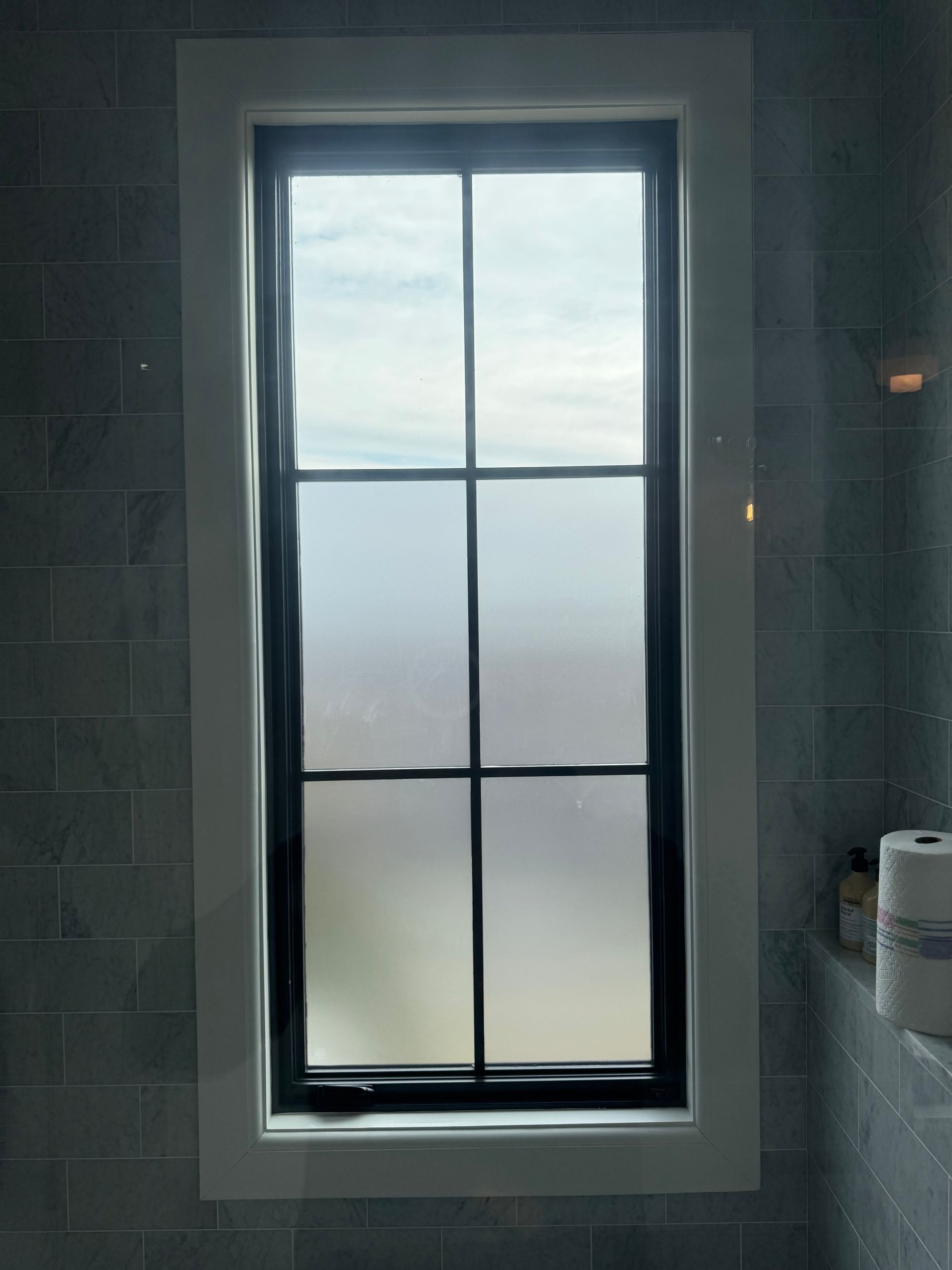 A tall window with a black frame and white trim set into a blue-gray tiled bathroom wall.