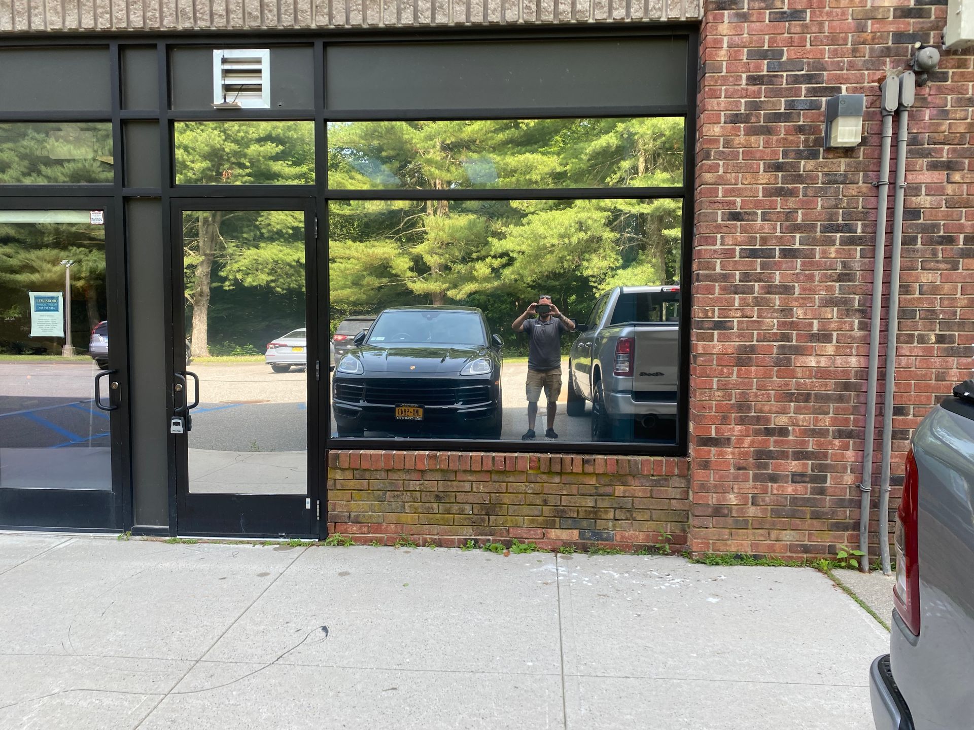 A person taking a photo of their reflection in a storefront window, with parked cars visible inside the reflection.