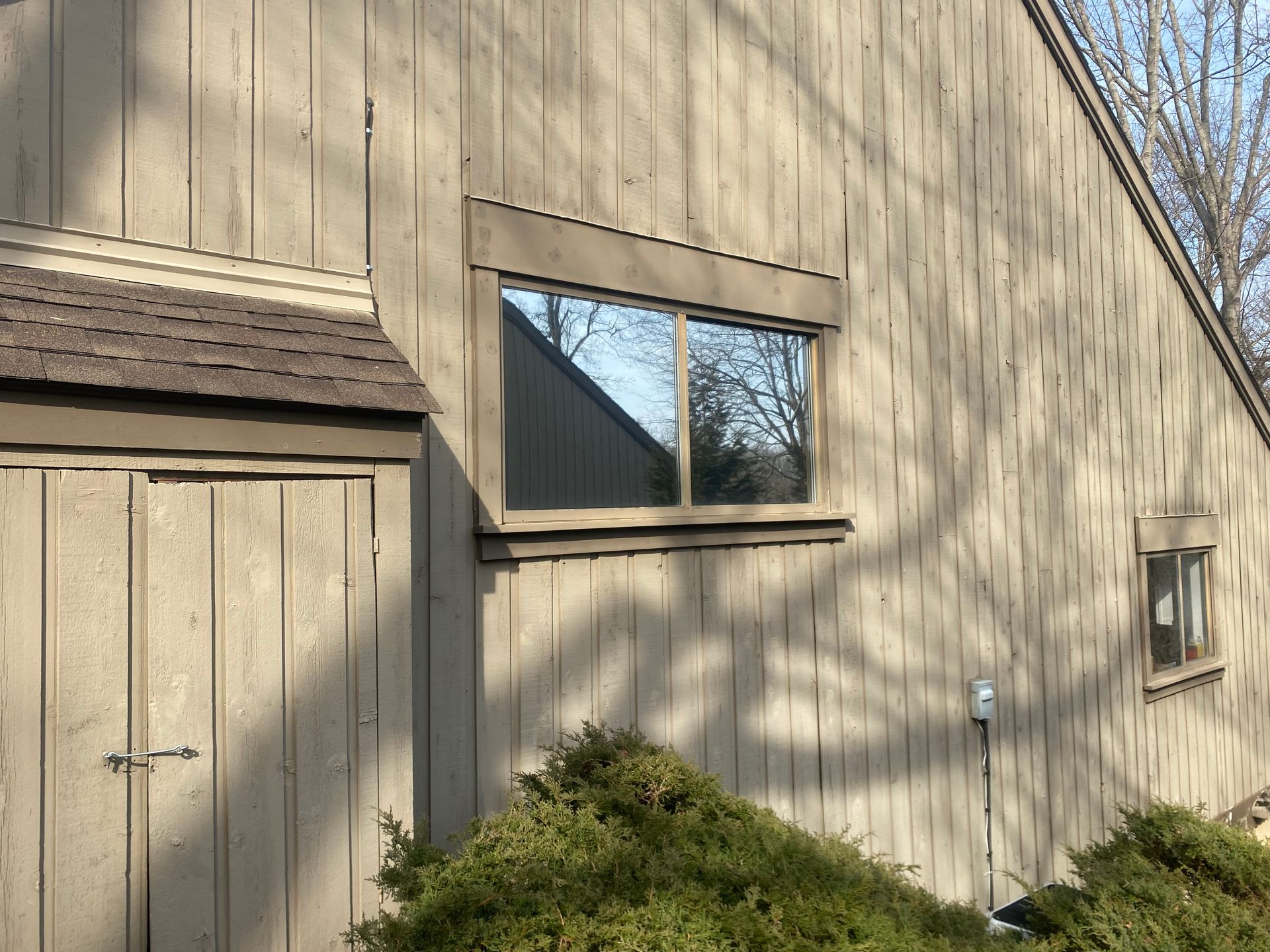 A tan-colored wooden building with a side door, a rectangular window, and tree shadows cast on the siding.